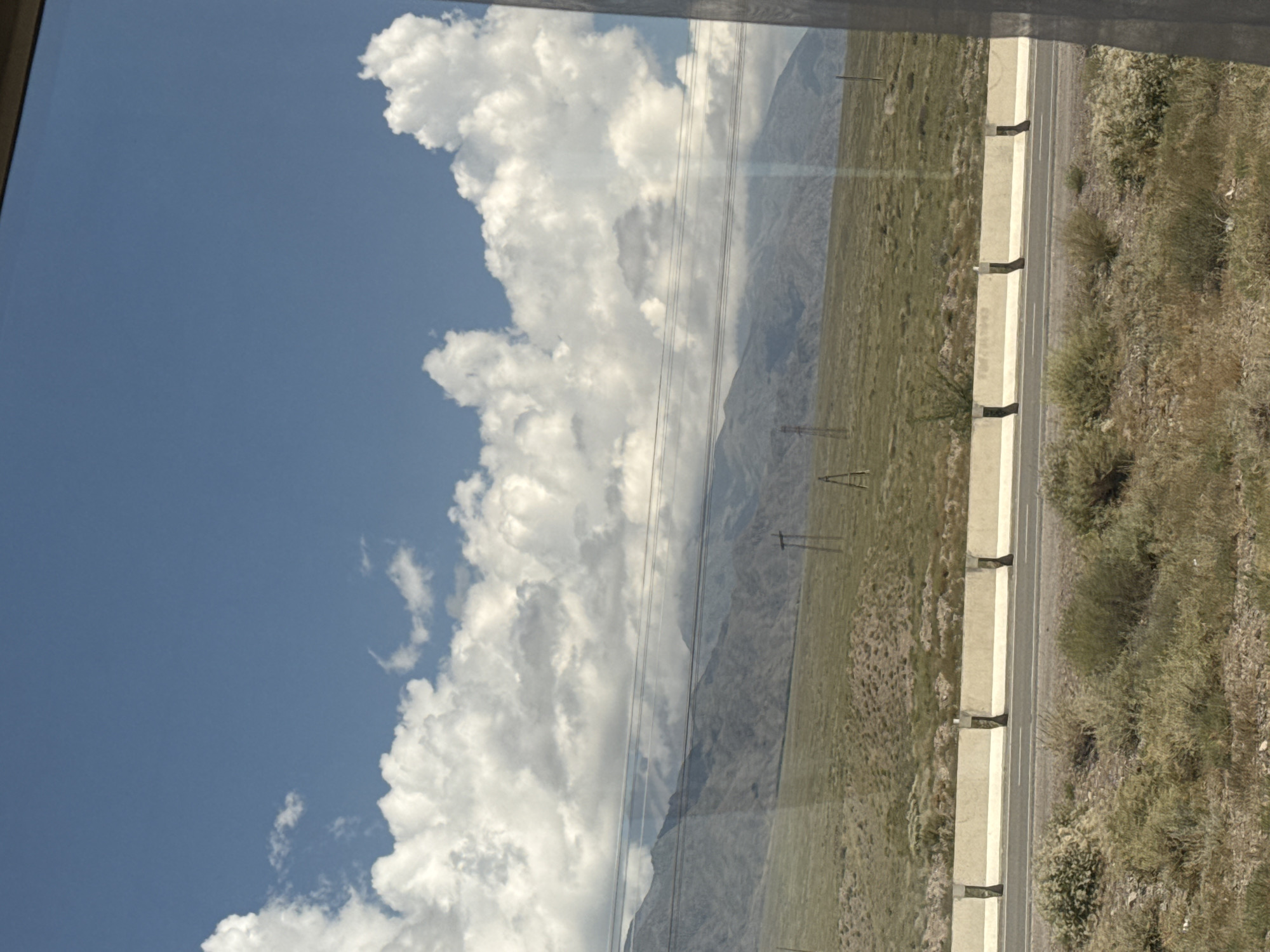 The image depicts a roadside scene in Kok-Moinok - 1, Kyrgyzstan. The foreground features dry, sparse vegetation and a highway with a concrete barrier. Beyond the highway, a grassy hillside rises towards a mountain range in the background, partially obscured by clouds. Power lines crisscross the scene horizontally, and several utility poles are visible on the hillside. The sky is blue with scattered cumulus clouds. No people or vehicles are present in the image. The overall impression is one of a vast, open landscape under a bright sky.
