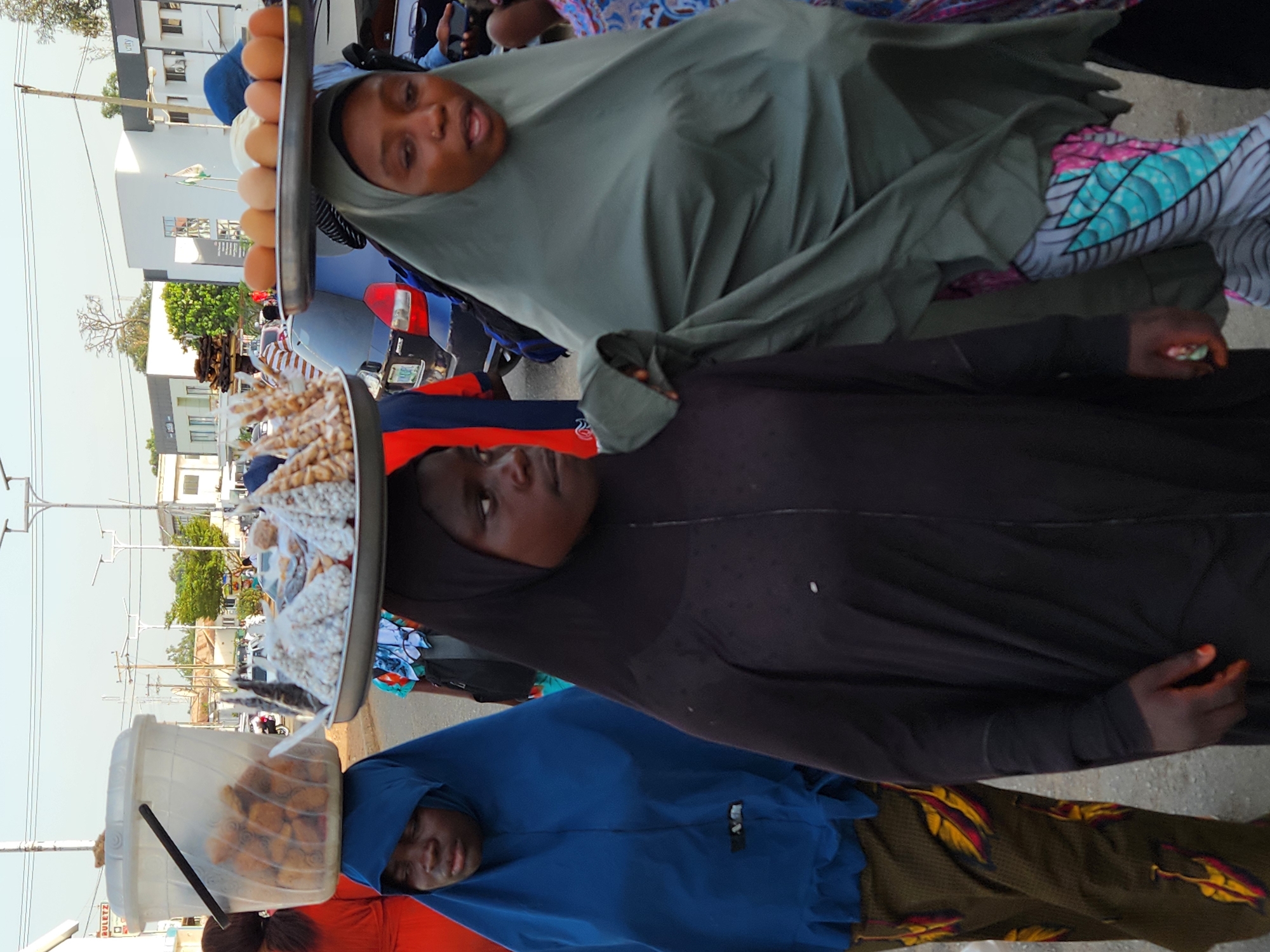 Three women in Jos, Nigeria are carrying goods on their heads. The first woman, at the top of the image, has a tray of eggs on her head and is wearing a green head covering. The second woman has a tray of what appear to be packaged snacks on her head, and she is wearing a black head covering. The third woman, at the bottom of the image, has a bucket of what look like fried snacks on her head and is wearing a blue head covering. The background shows a city street with buildings, power lines, and vehicles.