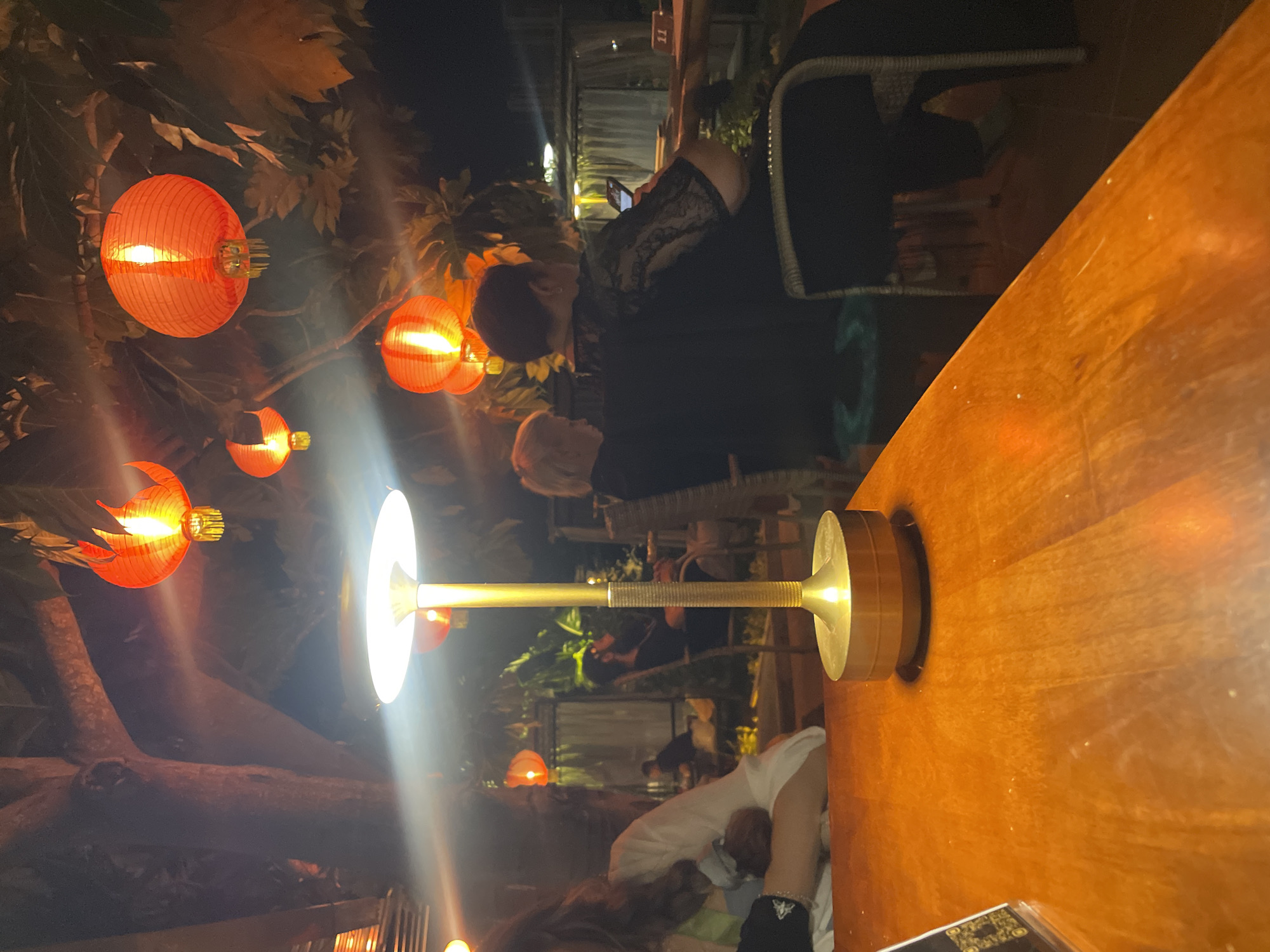 The image shows an outdoor setting, likely a restaurant or patio, in Bac Natural Heritage Area Trang, Vietnam. Several red lanterns are visible, strung among trees, providing illumination. A brass lamp sits on a wooden table, with its light reflecting off the surface. People are seated at the table; one is looking at a phone and another is in a white shirt. The table appears to be near other patrons, suggesting a social or dining environment. The scene is captured at night, with the lanterns and lamp contributing to the lighting.