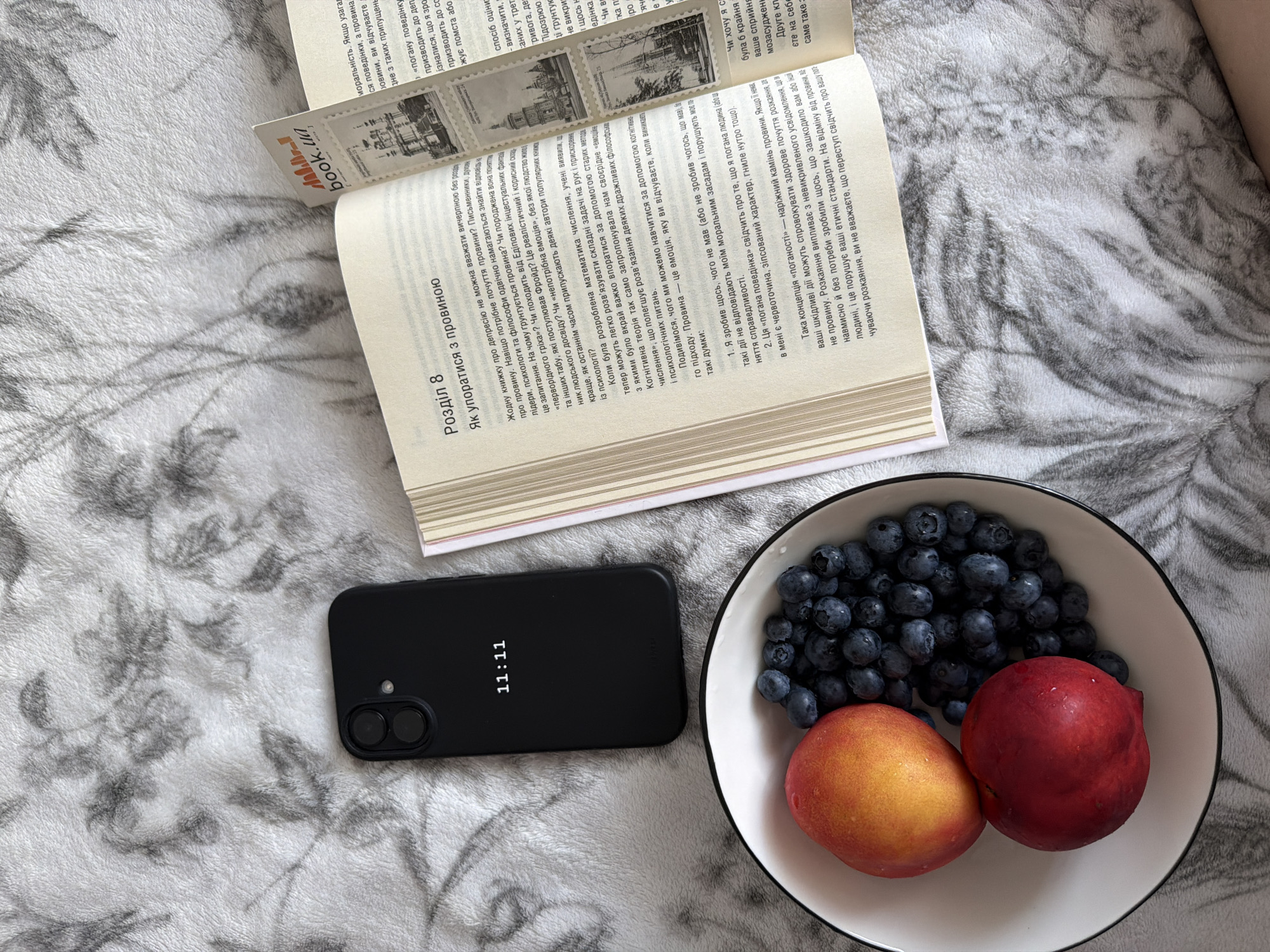 A black iPhone with the display showing "11:11" is placed on a textured gray blanket with a floral pattern. To the left of the phone is a white bowl filled with blueberries and two peaches. To the right of the phone is an open book with Ukrainian text and a bookmark from "book.ua" sticking out. The book appears to be on the same blanket. The scene is a flat lay, taken from directly above. There is no indication of a specific location in Kyiv, Ukraine.