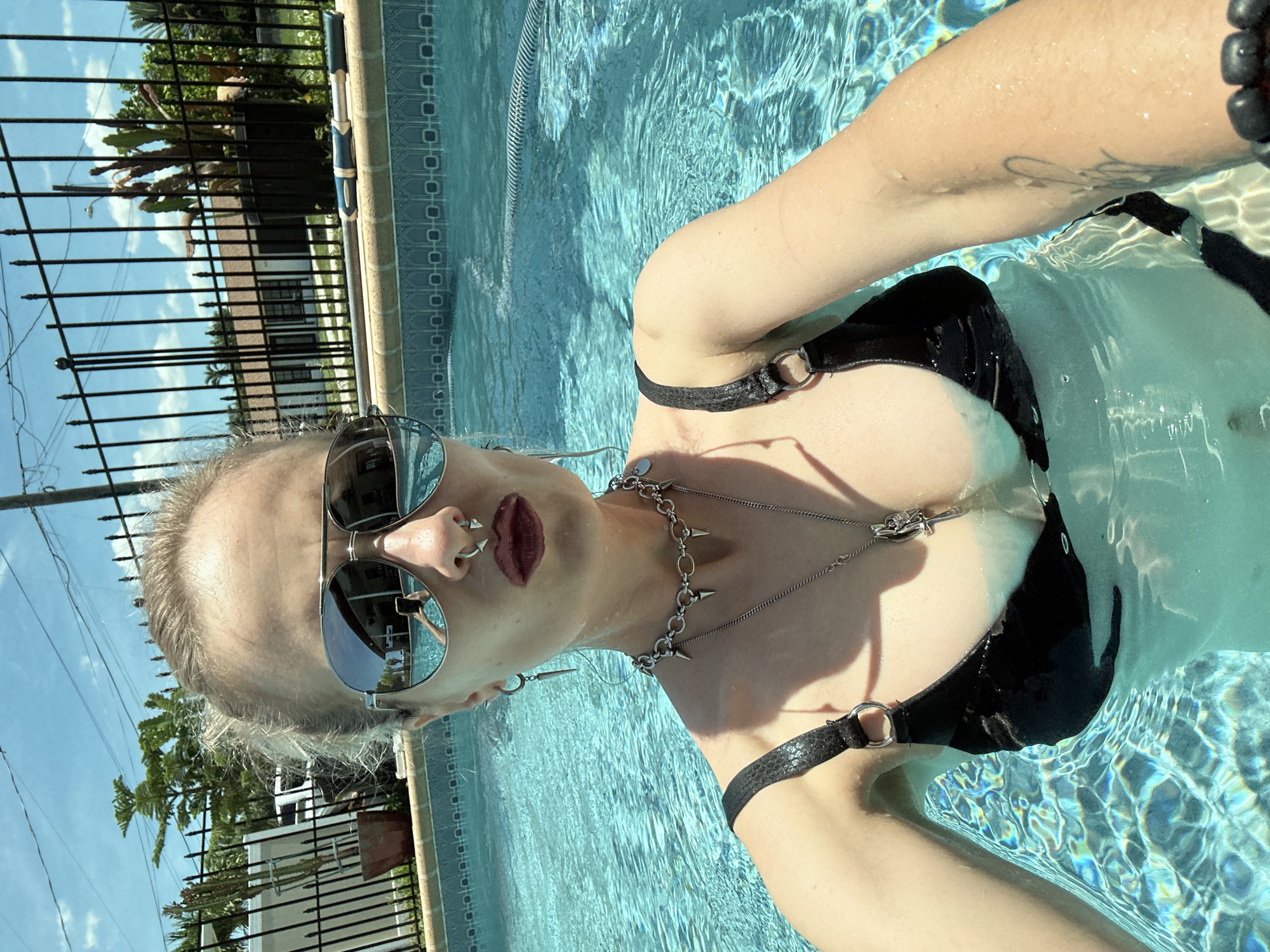 A woman with blonde hair and a nose ring is taking a selfie in a swimming pool on a sunny day. She is wearing a black bikini top with silver accents and several silver necklaces. Her sunglasses reflect the surroundings, which include a fence, a house, and trees. The pool water is clear and blue. A portion of a white building and a black metal fence are visible in the background, along with a plant in a pot. The scene suggests a residential setting in Cape Coral, Florida.