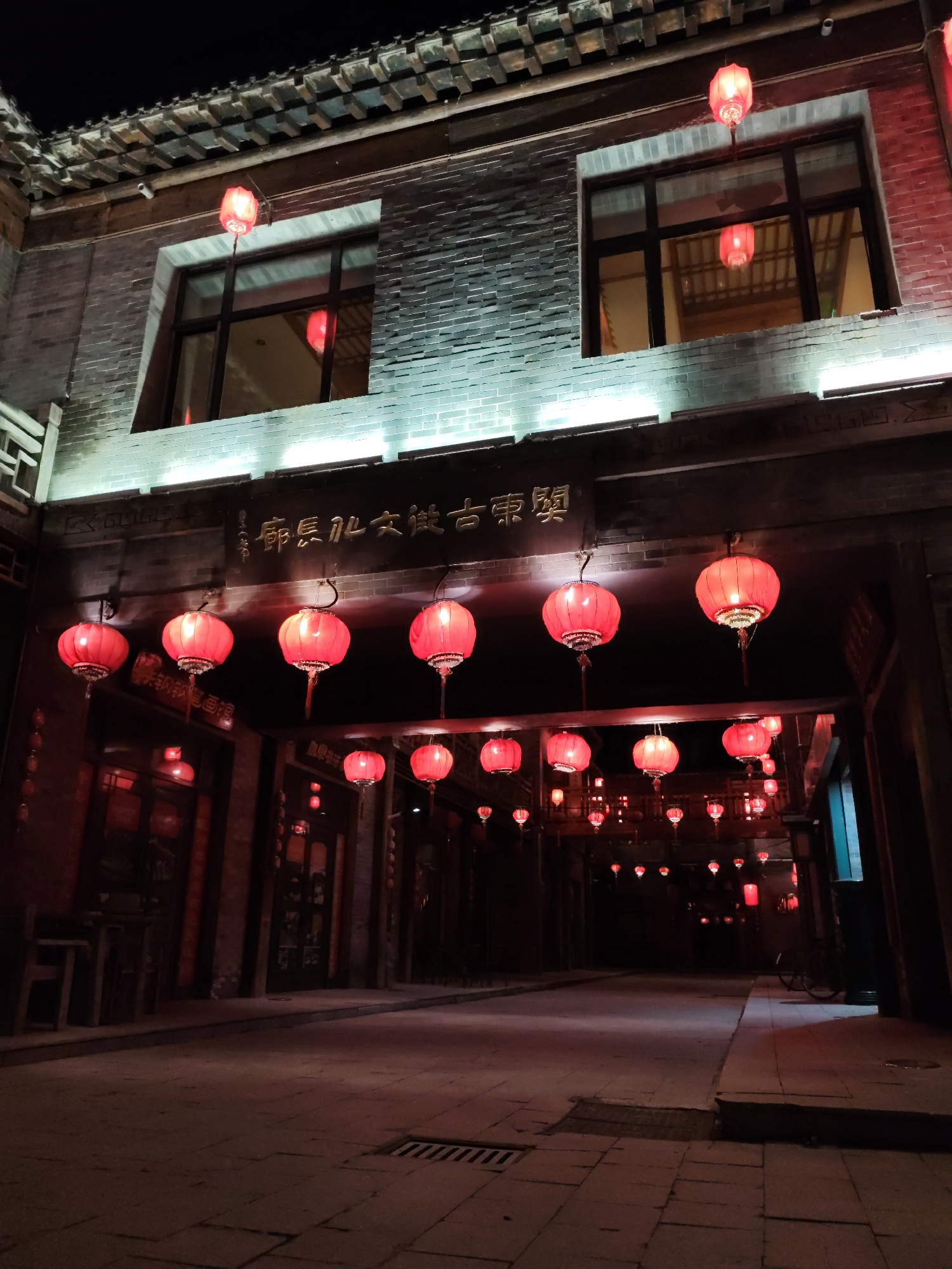 An exterior shot at night captures an alleyway decorated with numerous red lanterns. The lanterns hang from strings and are illuminated, casting a warm glow on the scene. The alleyway is lined with traditional Chinese architecture, featuring brick walls and ornate rooftops. The ground is paved with stone, and there are some tables and chairs visible on the left side of the alley. The overall atmosphere is festive and inviting, suggesting a cultural or historical district. The location is identified as Lindian, China. There are no people visible in the image.