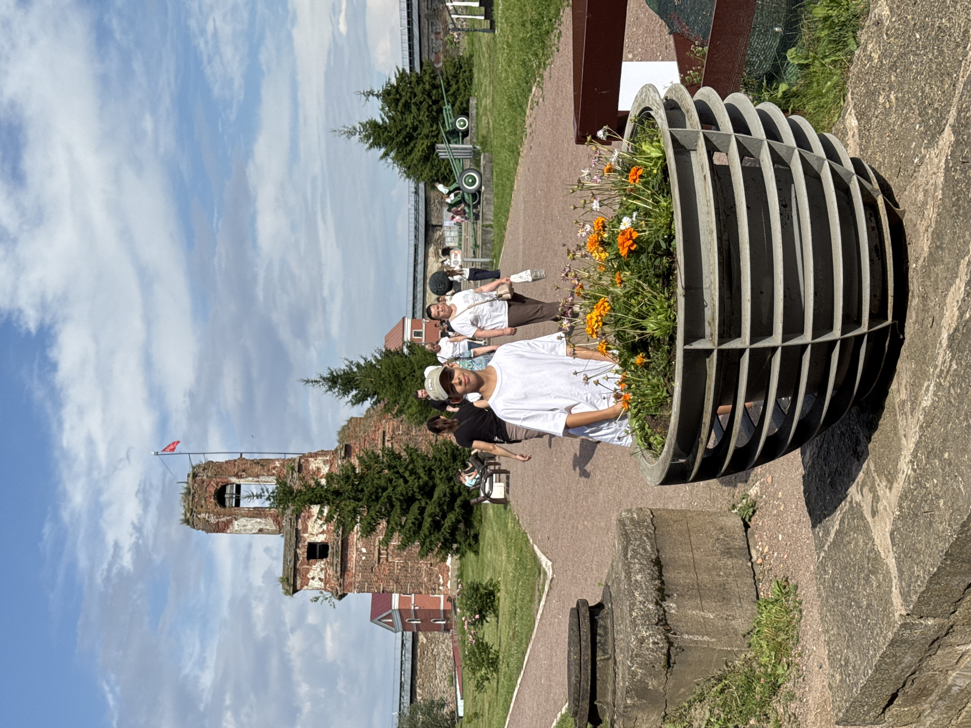 The image shows a boy in a white t-shirt and a cap standing in front of a large, decorative planter filled with orange and white flowers. In the background, there are several people walking on a path. To the left of the path, there is a red building and a ruined brick structure with a flag on top. To the right, there are evergreen trees, and further in the distance, there is a display of artillery. The scene appears to be a park or historical site in Shlisselburg, Russia, possibly Oreshek Fortress, given the architectural style and the presence of historical military equipment. The sky is blue with scattered clouds, and the sun is shining, casting shadows. The ground is a mix of gravel paths and grass.