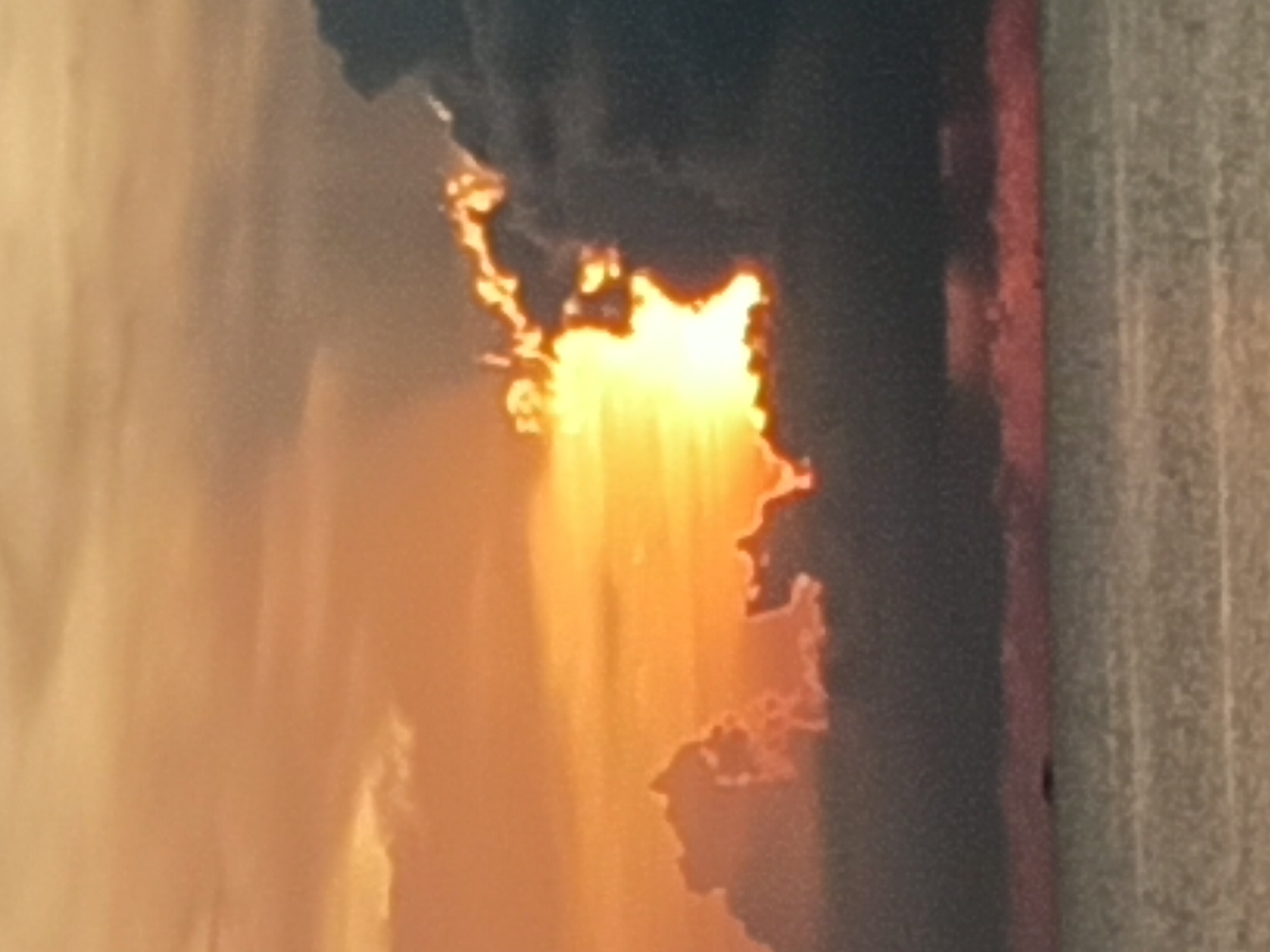 The image shows a close-up view of a fire burning, with flames emanating from a dark area. The fire is bright orange and yellow, contrasting with the surrounding darkness. To the right of the flames, there is a vertical structure, possibly a wall or part of a building. There are no people visible in the image. The location of Baan Wok Tum, Thailand is not discernible from the image.