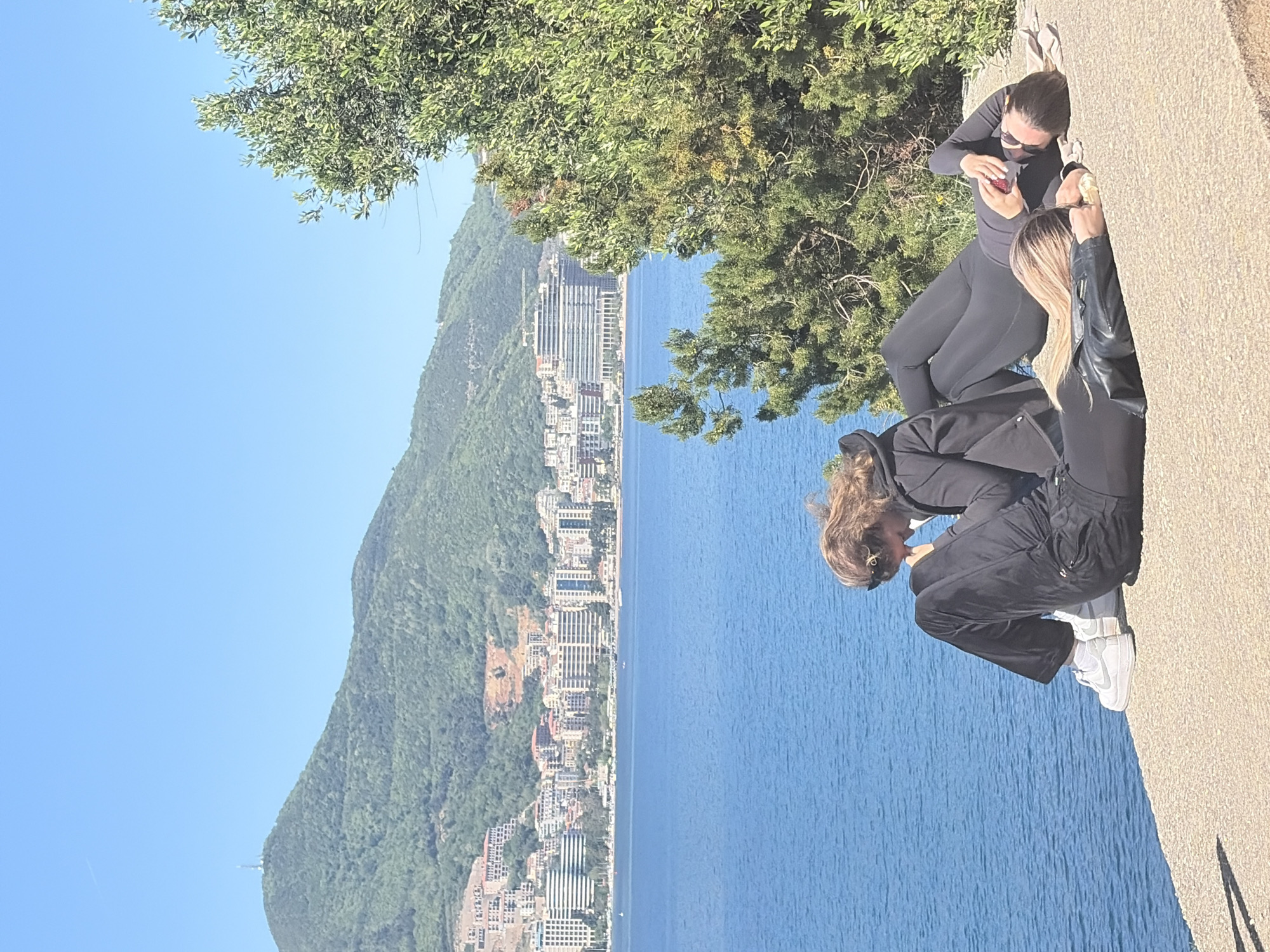 Three women are standing on a beige concrete ledge overlooking the city of Budva, Montenegro, located on the Adriatic Sea. A hillside covered with greenery stretches down to the coastal city with multi-story buildings. The woman in the foreground is kneeling and appears to be looking at something. The woman in the middle is standing up, dressed in black, and has long blonde hair. The woman behind her is standing up and appears to be eating a strawberry. The weather is clear and sunny.