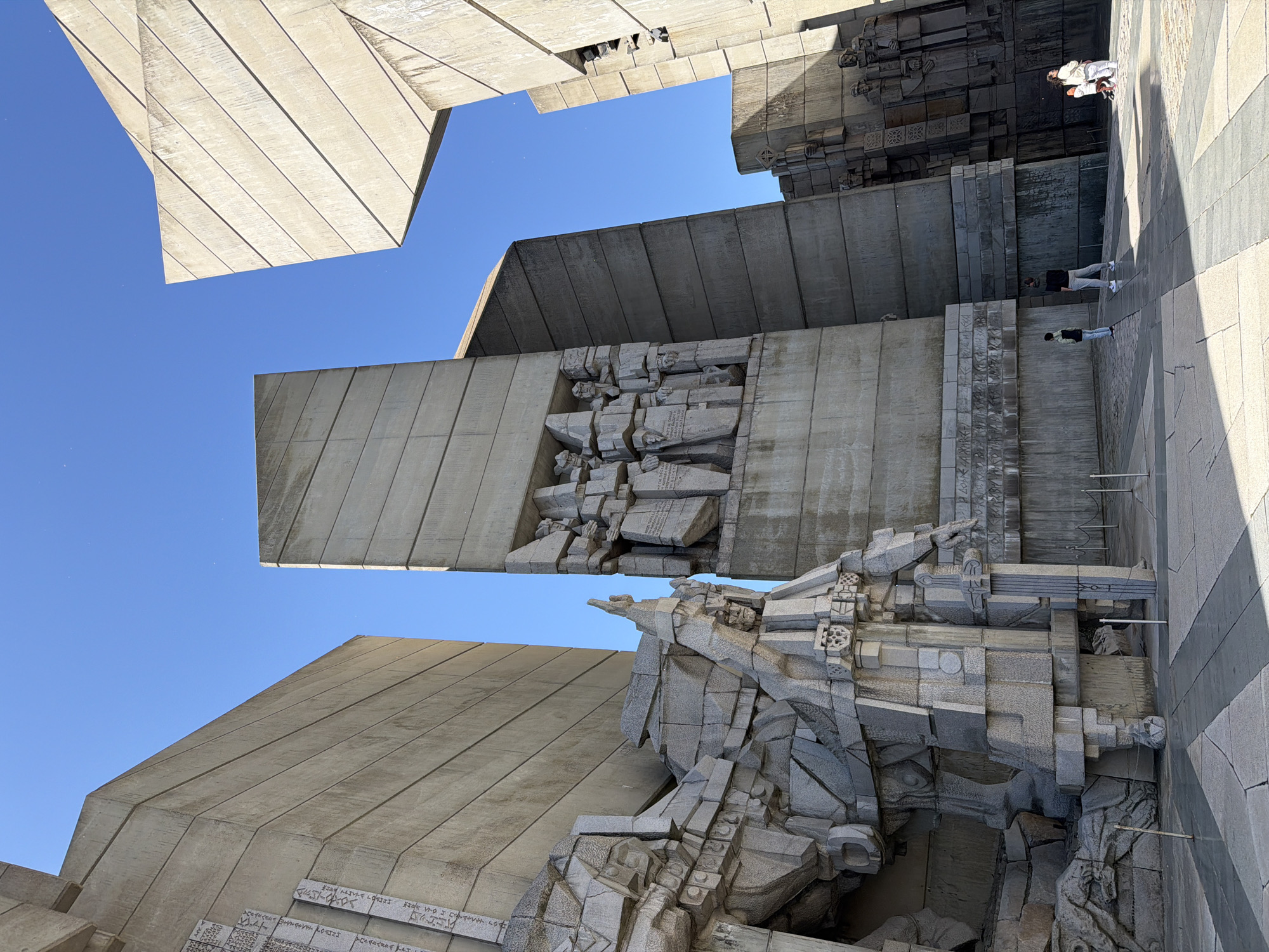 The image captures a low-angle view of the Founders of the Bulgarian State Monument, located in Shumen, Bulgaria. The monument is constructed from large, angular concrete blocks.
The sky is clear and blue. The monument is composed of various geometric shapes, including rectangular slabs and fragmented sculptural elements. Some slabs have inscriptions on them. The sculptures appear to be in a Cubist style.
Three people are visible in the upper right part of the image, standing on a paved surface. The pavement is made up of light and dark gray tiles arranged in stripes.