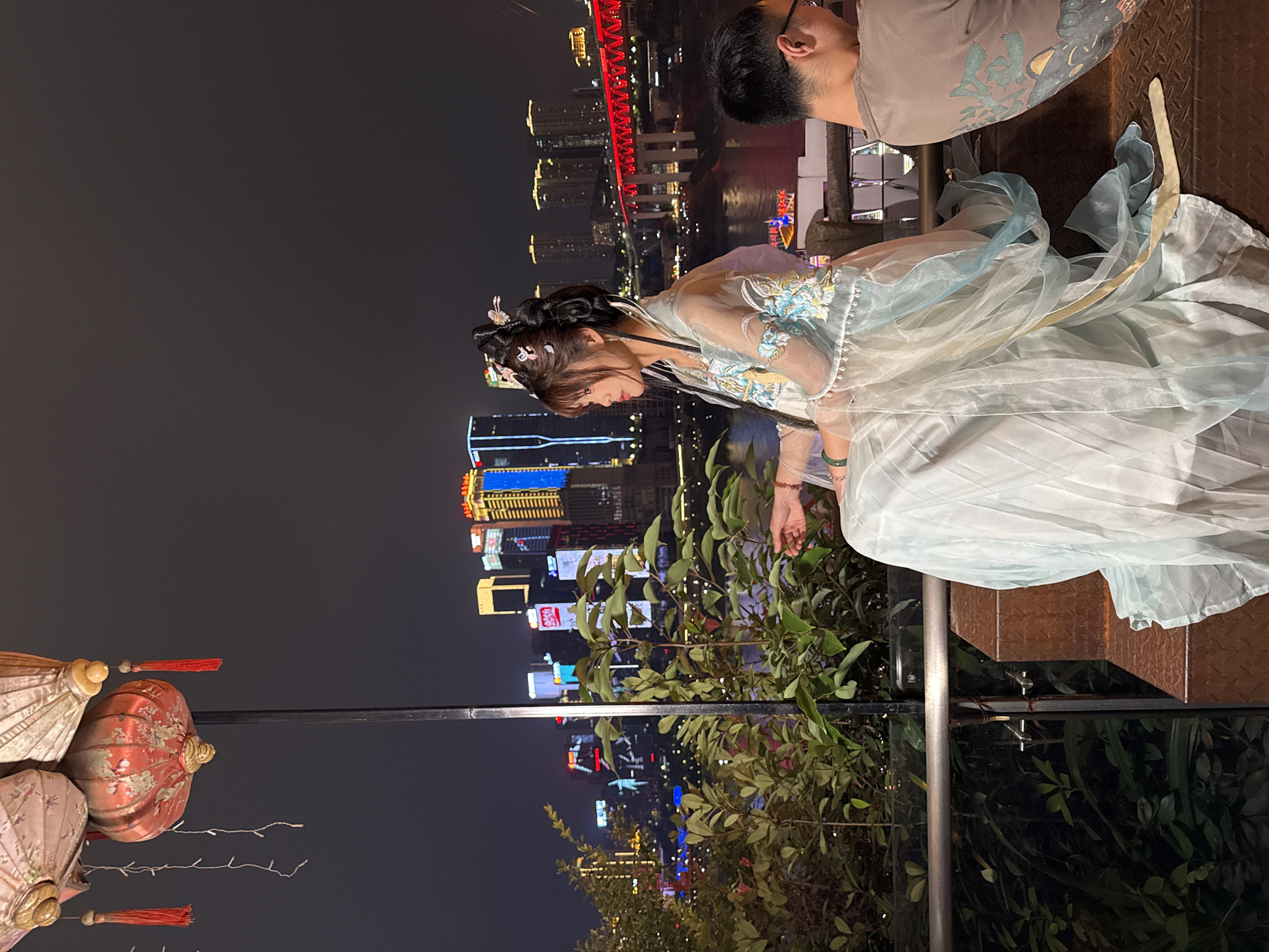 A horizontally-oriented medium shot at night showcases a woman in traditional Chinese clothing. She has dark hair styled with traditional ornaments, and wears a light-colored dress with sheer layers and embroidered details. Her head is bowed, and her hands rest near green leafy foliage.
Behind her is a nighttime cityscape. A tall red bridge is visible, reflecting in the water. City buildings light up the background.
Next to the woman is a man whose shoulder and head are visible. He wears glasses.
In the left foreground are Chinese lanterns.