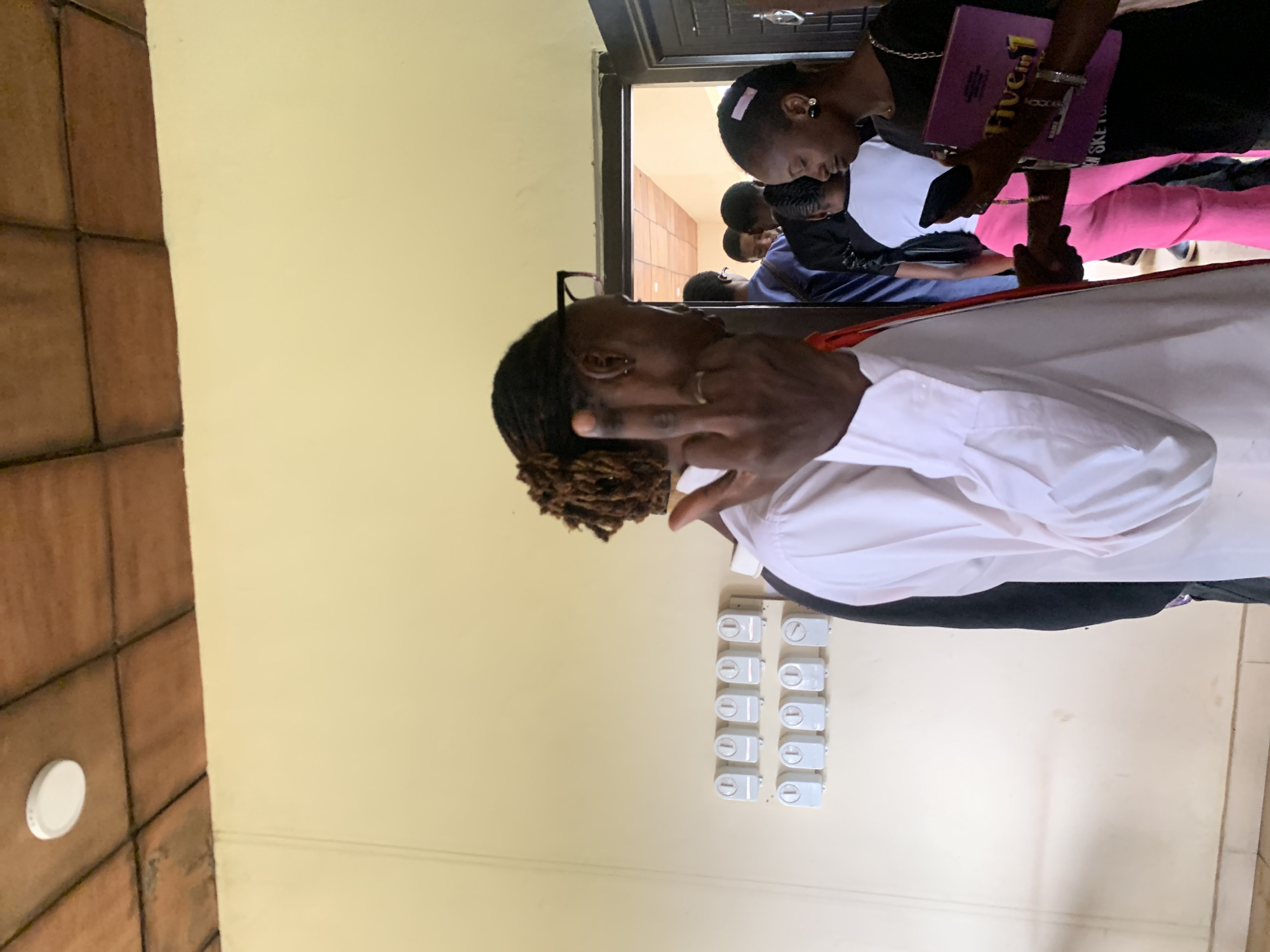 The image shows an indoor scene with several people. In the foreground, a person wearing a white collared shirt is facing away from the camera, with their right hand raised in a gesture. This person has dreadlocks and is wearing glasses and a ring on their finger. To the right, a woman in a black top is looking down at a purple folder in her hands, which appears to be a "Five in 1" book. She has a pink ribbon in her hair and is wearing earrings and bracelets. Behind them, several other people are visible, including some standing near an open doorway that leads into another room with a tiled ceiling. On the wall to the left, there is a row of twelve circular electrical switches. The walls are painted a light yellow color, and the floor appears to be tiled. The setting is an indoor area, possibly a school or community center, in Farin Yashe, Nigeria.