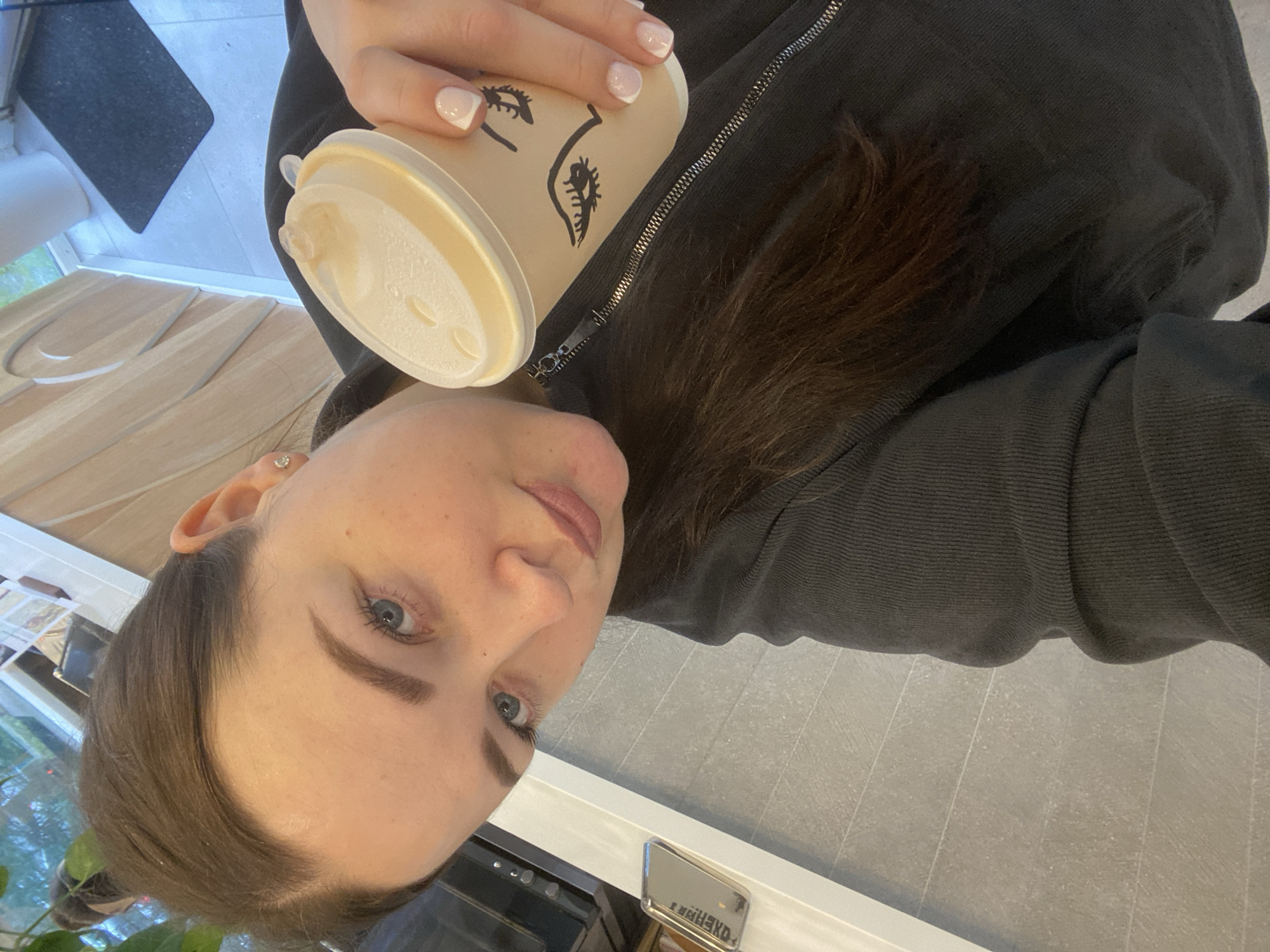 A woman is holding a disposable coffee cup with a cartoon face printed on it. She is wearing a gray hoodie and her nails are painted with a French manicure. She appears to be inside, possibly in a business in Sochi, Russia, based on the name tag on the counter behind her. The floor has gray tiles.