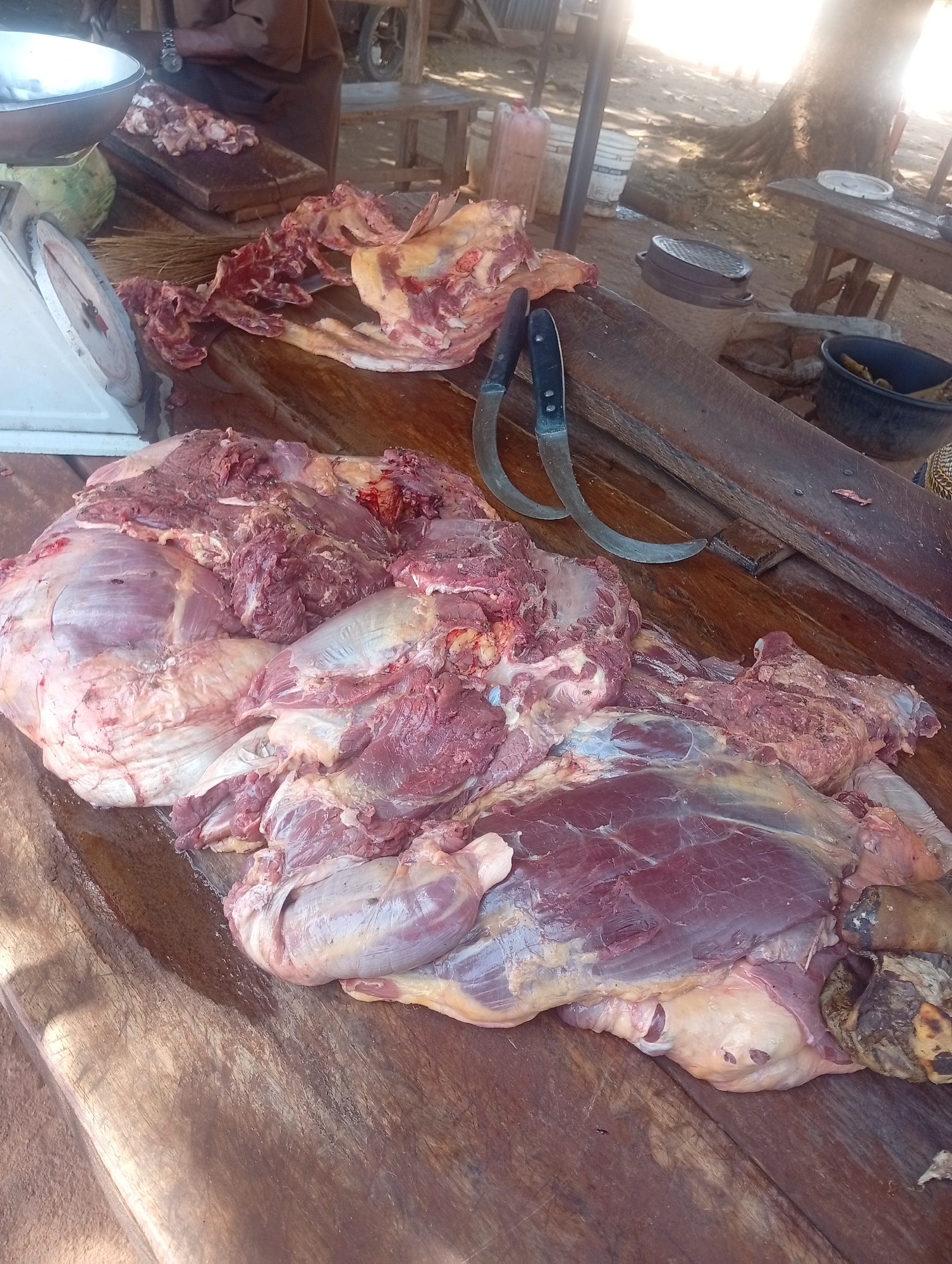 The image shows a large amount of raw meat on a wooden table. Two curved knives with black handles are placed next to the meat. In the background, there is a scale, more meat on a cutting board, and various containers. The location appears to be an outdoor market or butcher shop in an unknown city.