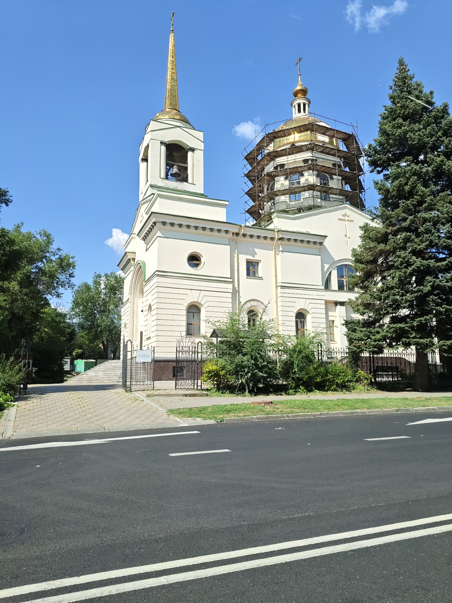 The image depicts the Church of the Nativity of the Most Holy Mother of God, located in Rostov-on-Don, Russia. The church is a prominent religious building with a light-colored facade. A tall, golden spire, topped with a cross, rises from a bell tower. Adjacent to the bell tower is a large, golden dome, partially covered in scaffolding, indicating ongoing renovation or construction work. The church is surrounded by lush green trees, and a paved pathway leads to its entrance. In the foreground, a dark asphalt road with white lane markings runs across the scene. The sky above is a clear blue with a few scattered white clouds.