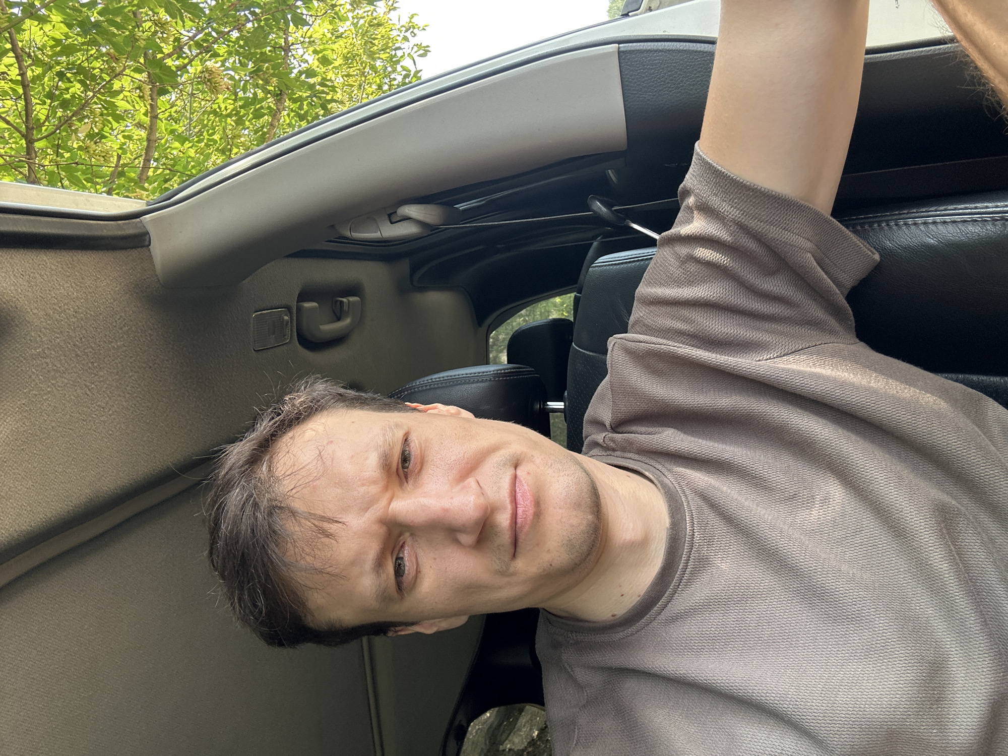 The image shows a medium shot of a man sitting in the driver's seat of a vehicle. He has brown hair, fair skin, and is wearing a brown t-shirt. His facial expression appears serious or concerned, with his eyebrows furrowed and his mouth slightly downturned. The interior of the car is visible, including a black leather seat and the car's ceiling. Outside the car, green foliage and trees are visible, suggesting an outdoor setting. The lighting indicates it is daytime. The image does not provide any specific information about the location in the city of Makiv, Ukraine.
