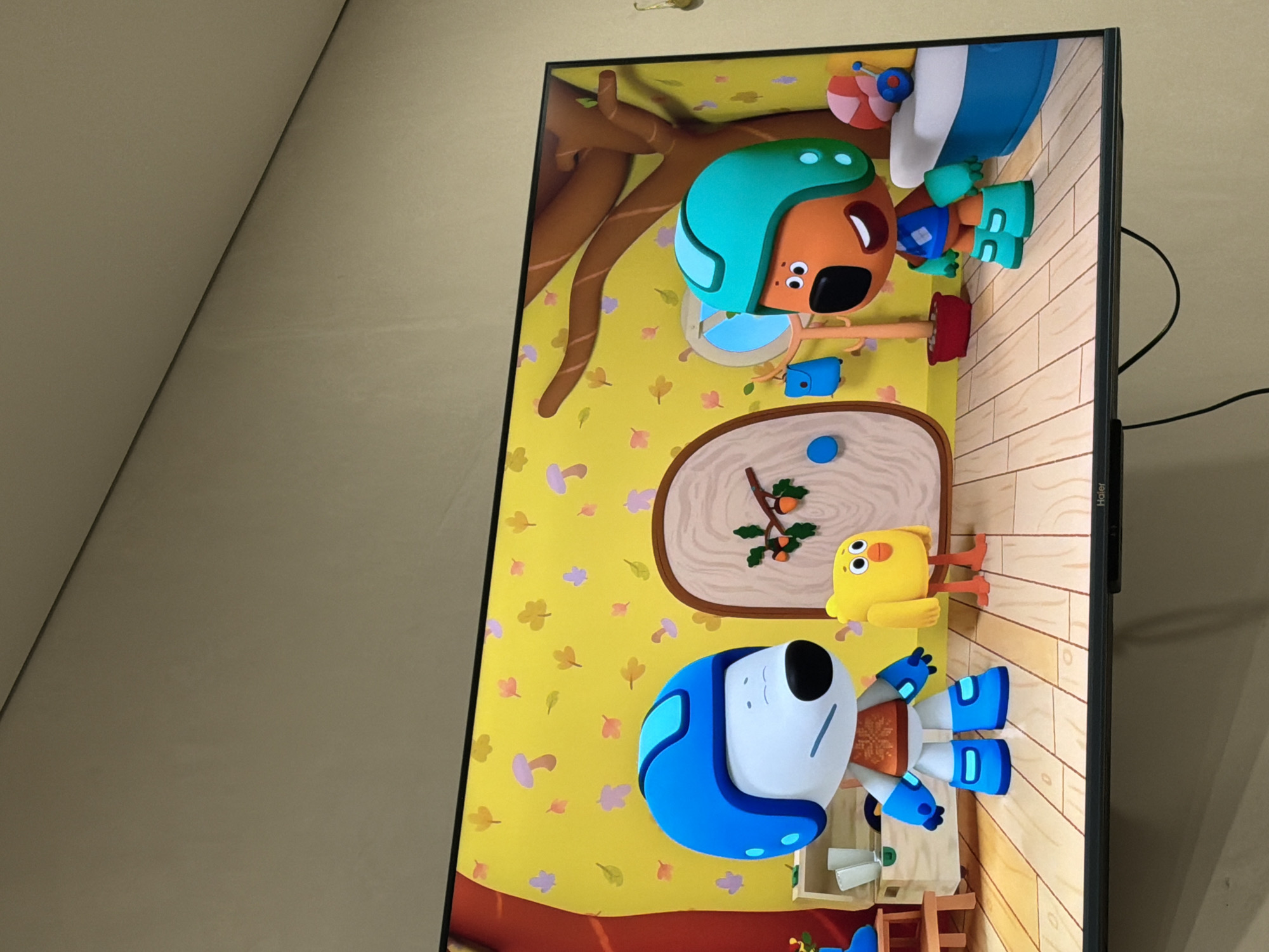 The media file shows a television screen displaying a cartoon scene. The scene features several animated characters, including a brown dog wearing a turquoise helmet and blue shorts, and a yellow bird with orange legs. Another character, a white robot with blue accents and a blue helmet, is also visible. The background is a bright yellow wall decorated with colorful autumn leaves and mushrooms. There is a wooden floor and a tree with brown branches. A small blue mouse and a red pot are also present. The cartoon appears to be aimed at children. The television set itself is black and has a "Haier" logo on the bottom right side. The overall mood of the scene is cheerful and playful. The image is taken in Krasnyi Krym, Russia. There is no discernible time of day or weather information, and no specific location cues beyond the country.