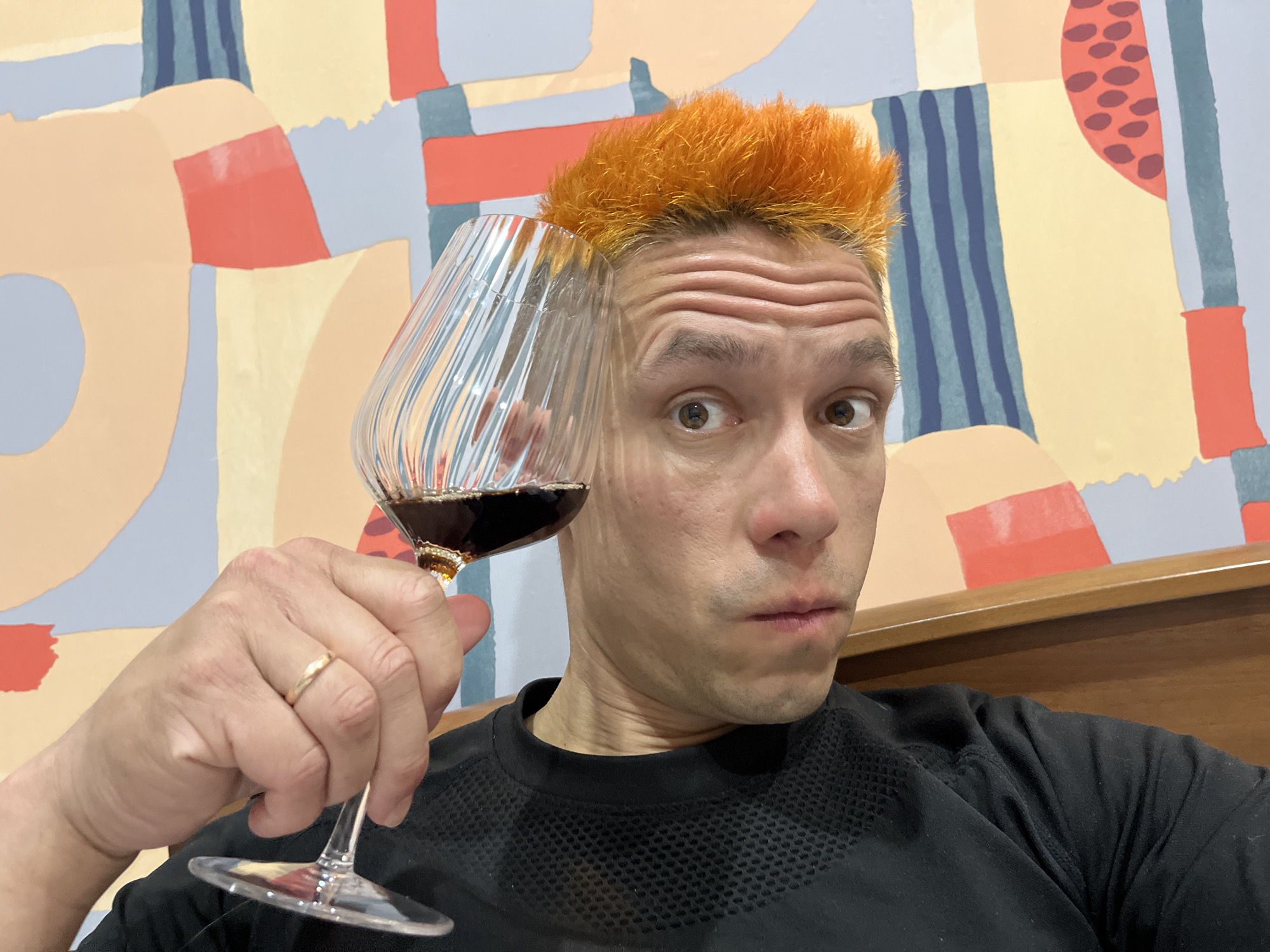 A fair-skinned person with orange hair holds a glass of red wine, partially obscuring their face. The person wears a black shirt and a gold ring on their left ring finger. The background features a colorful abstract painting with red, yellow, blue, and grey sections. The photo was likely taken indoors. There is no information in the image that confirms the location as Chaikovsky, Russia.