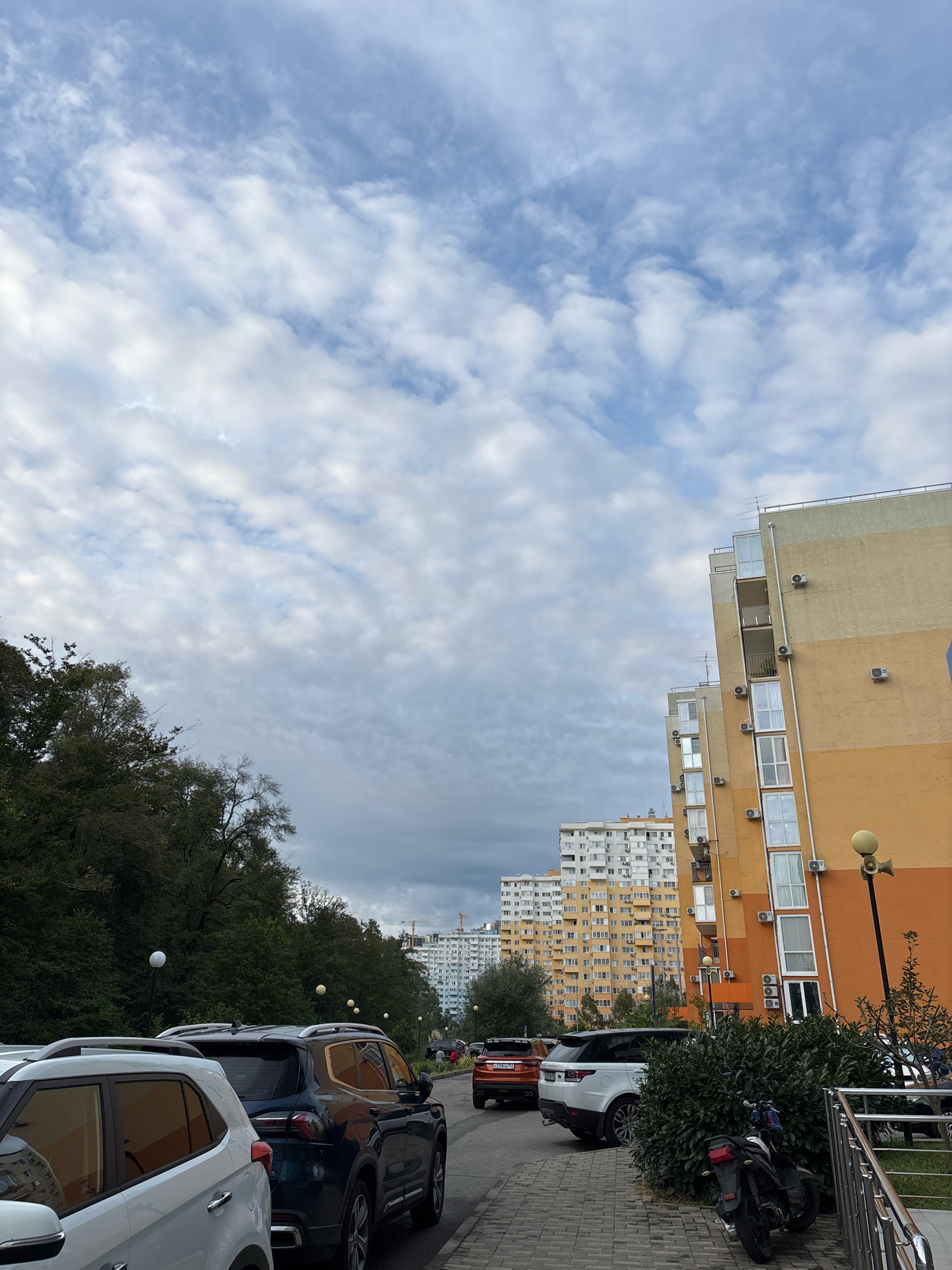 The image shows a street scene with several parked cars, including a white SUV in the foreground on the left, a dark blue SUV, and an orange SUV. Behind the parked cars, a multi-story apartment building with an orange and yellow facade dominates the right side of the frame, featuring numerous balconies and air conditioning units. Further in the background, more apartment buildings are visible, some appearing older and painted white. Trees line the left side of the street, and a sidewalk with a metal railing is present in the foreground on the right. A motorcycle is parked near the sidewalk. The sky is partly cloudy with a mixture of blue sky and white clouds. The overall impression is of a residential area in an urban or suburban environment.