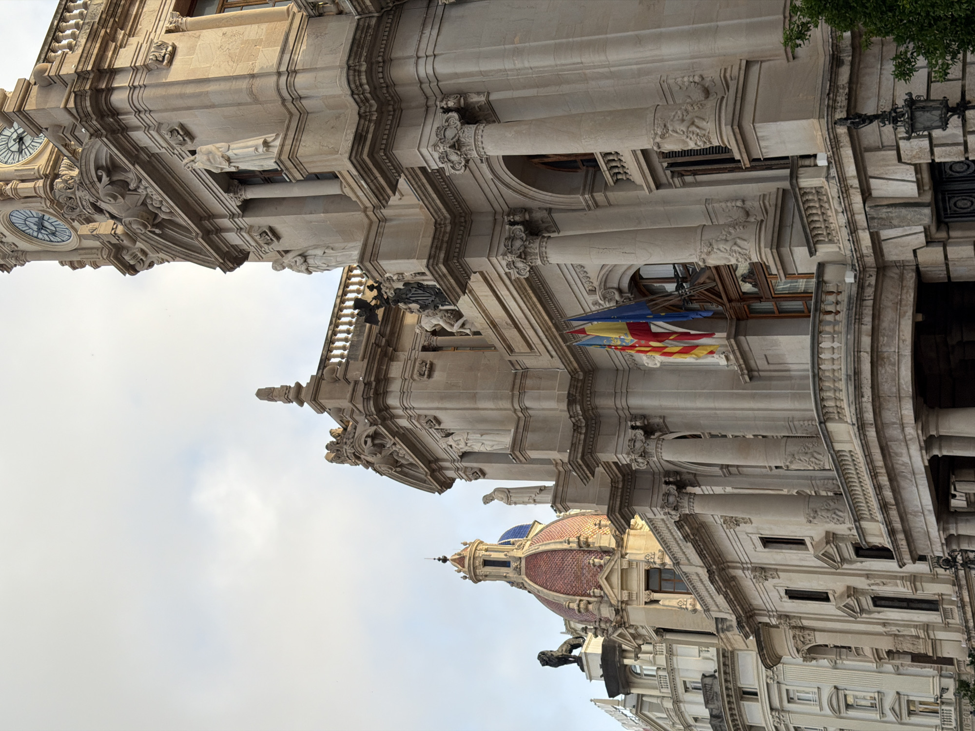 The photograph captures a low-angle, upward view of the facade of a grand, ornate building, likely a government or historical structure. The architecture is a mix of neoclassical and baroque styles, characterized by extensive stonework, intricate carvings, columns, and statues.
Dominating the right side of the frame is a tall, imposing tower with a large clock face visible at the top, featuring Roman numerals. Below the clock, decorative carvings and multiple statues of robed figures are integrated into the facade. The building material appears to be a light-colored stone, possibly sandstone or limestone, showing signs of age and weathering.
To the left of the main tower, a domed structure with a red-tiled roof and a blue-tiled accent is visible. A dark statue of a lion stands guard on a pedestal in front of this dome. Another, more modern-looking building with a pale facade and numerous windows is partially visible behind and to the left of the domed structure.
Flags are mounted on poles attached to the facade near the center of the image. These appear to be regional or national flags, with red, yellow, and blue colors prominent. The sky above is overcast with some patches of lighter clouds. The overall impression is one of historical grandeur and architectural complexity.