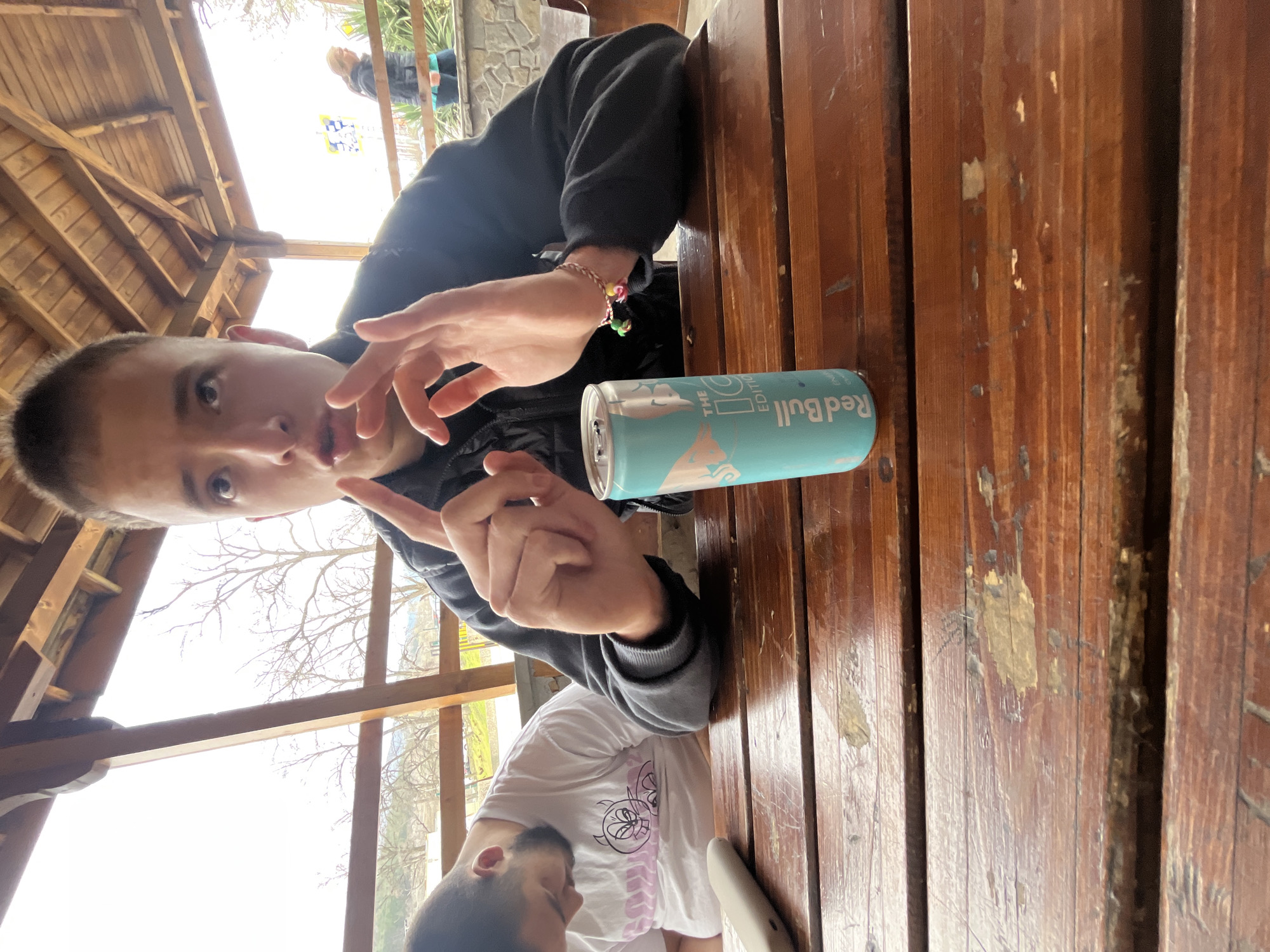 A selfie-style photograph shows a young person seated at a wooden table. A can of Red Bull is placed to their right on the table. The person is wearing a black jacket and has a beaded bracelet on their wrist. The person is looking at the camera, with fingers raised in a V-shape. Another person is visible in the lower part of the frame, wearing a white t-shirt. The photograph appears to have been taken in an outdoor seating area with a wooden structure overhead, possibly a shelter or gazebo. In the background, trees and a building are visible through the structure's openings. The location is in Asenovgrad, Bulgaria.