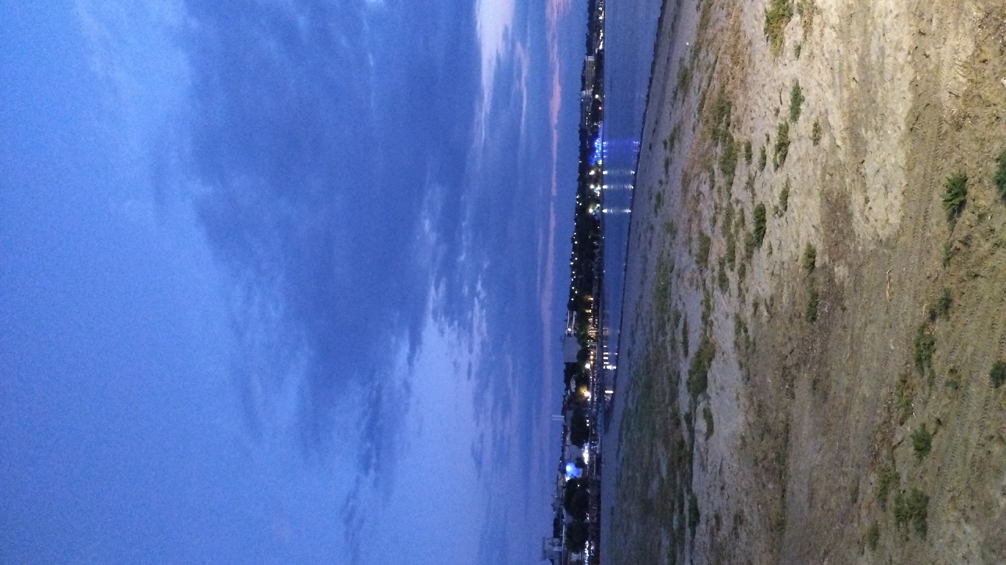 A twilight scene shows a beach in Neoi Epivates, Greece, with the Aegean Sea in the background. The sky is a deep blue with scattered clouds. Along the coastline, buildings and streetlights are illuminated, casting reflections on the calm water. The foreground consists of sand with patches of sparse vegetation. The image depicts a peaceful, serene coastal environment at dusk.