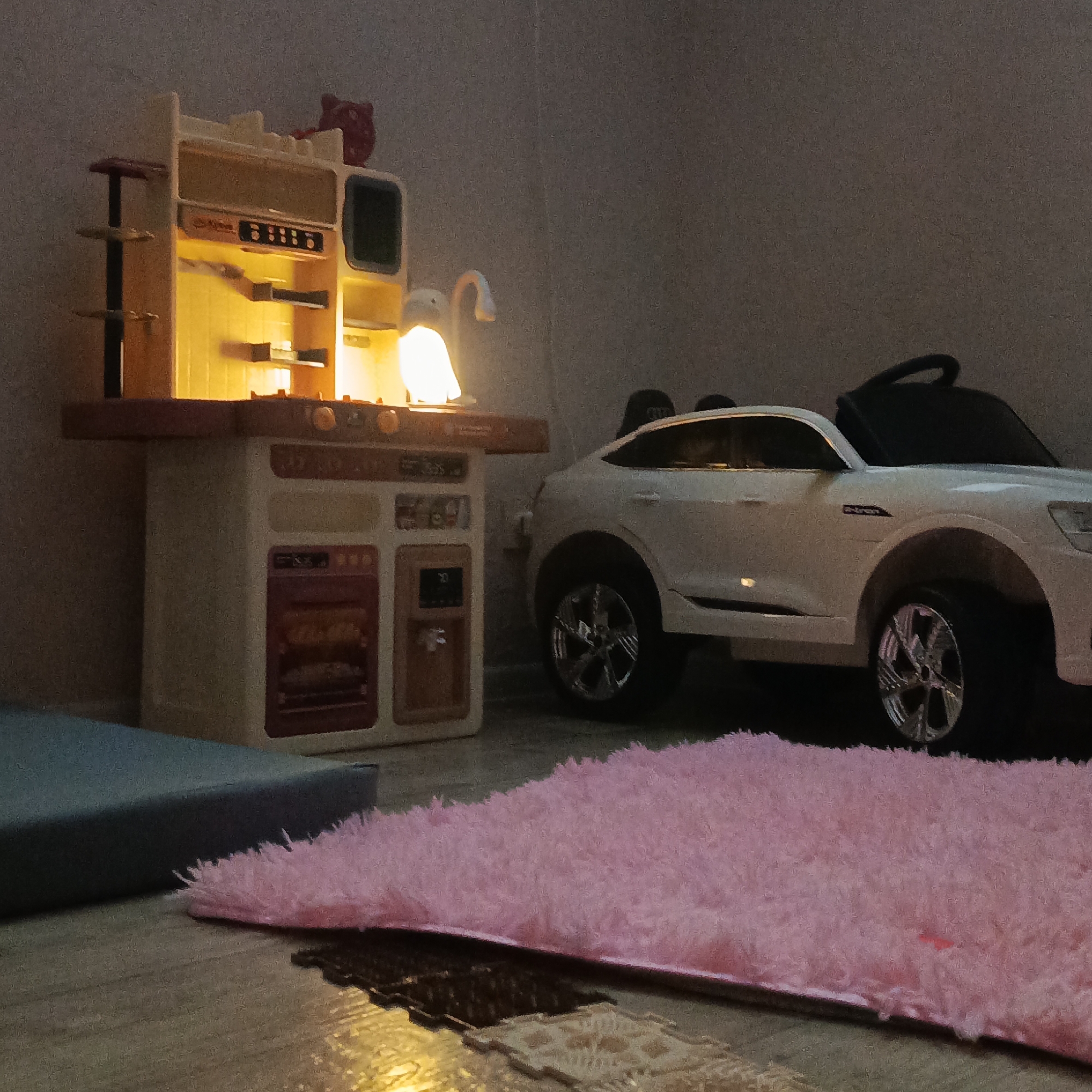 A low-light indoor scene shows a children's play area in Марковский, Russia. A toy kitchen, illuminated from within, stands to the left. A white toy car is positioned next to the kitchen. A pink fuzzy rug is in the foreground, partially covering a beige patterned rug. A blue cushion sits to the left of the pink rug. The background is a plain wall. No people are visible.