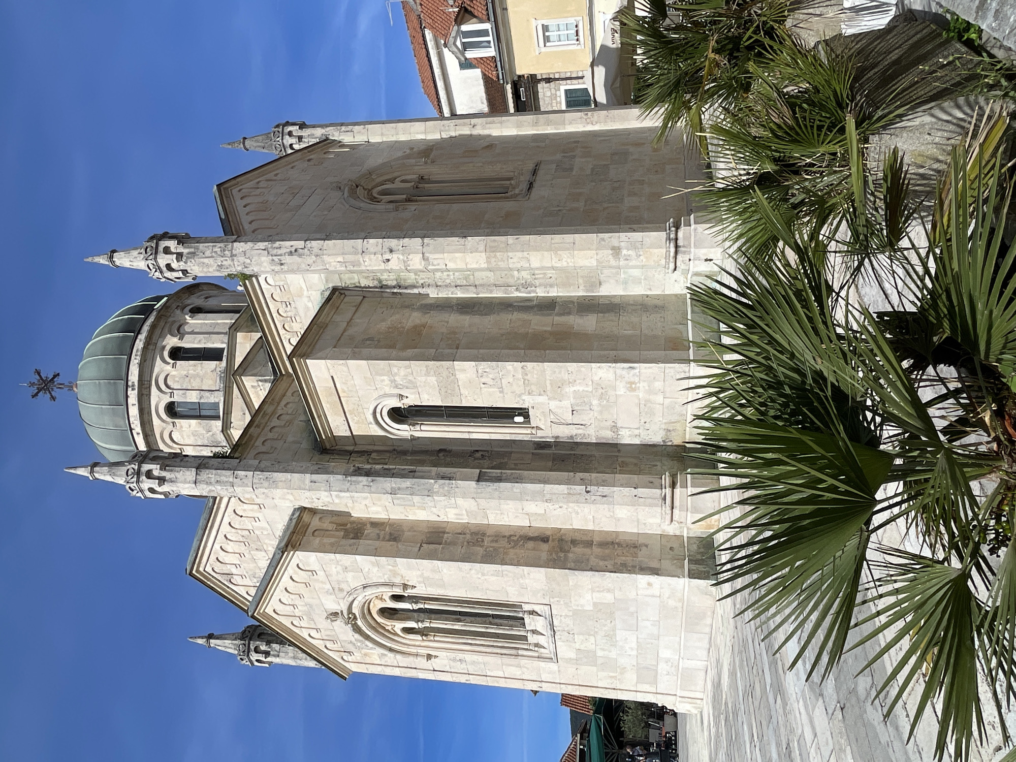 The image presents a prominent, ornate stone building, likely a church or cathedral, under a clear, vibrant blue sky. The building, constructed from light-colored, possibly limestone, blocks, features a distinguished greenish-grey dome topped with a cross-like finial. Several pointed spires and decorative finials adorn its roofline and corners. The facade is characterized by tall, arched windows with intricate stone tracery, along with carved details, cornices, and moldings, indicating a rich architectural style, possibly Mediterranean or classical.
To the right, and partially in the foreground, several large, green palm trees extend into the frame. Their fronds cast sharp, distinct shadows on the building and ground, suggesting bright, direct sunlight, likely from midday or early afternoon.
In the background, behind the main structure, glimpses of other buildings are visible. These buildings have traditional red-tiled roofs and facades in yellow and white colors. On one of these background buildings, a white awning displays faint, partially visible text that appears to be "EMIN".
The overall scene captures a serene, sun-drenched atmosphere, emphasizing the historical architecture and natural beauty of the setting. The ground visible at the bottom of the image appears to be paved. No people or specific activities are depicted, allowing the architectural and natural elements to dominate the composition. The combination of stone architecture and palm trees is consistent with the provided location context of Herceg Novi, Montenegro.