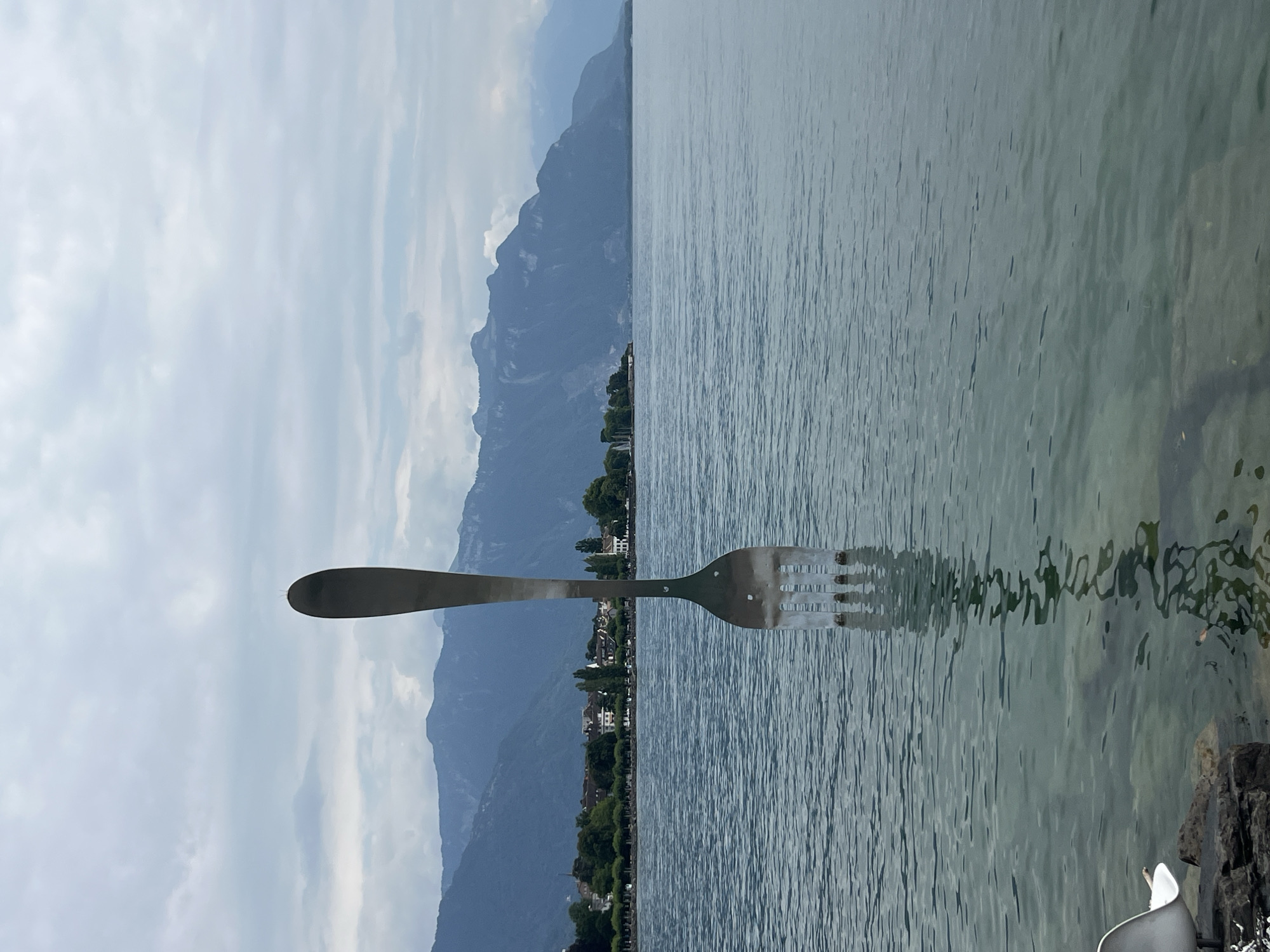 An oversized silver fork sculpture stands in the clear, shallow water of Lake Geneva, Vevey, Switzerland. The sculpture, positioned vertically with its tines submerged, is reflected in the rippling surface. In the background, a tree-lined promenade and buildings of Vevey are visible along the shore, backed by a range of hazy blue mountains under a cloudy sky. The foreground features dark, wet rocks and a portion of a white chair.