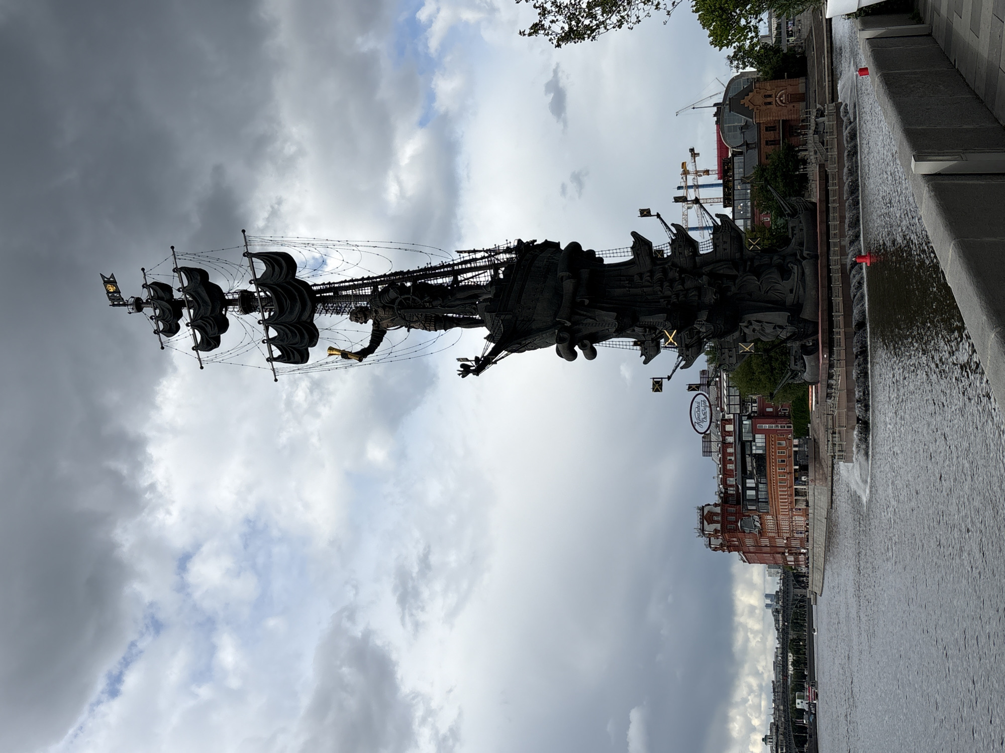 The image shows the Peter the Great Statue in Moscow, Russia, from a low-angle perspective. The statue depicts a ship with multiple sails and figures, set against a cloudy sky. Part of a river is visible in the foreground, with a fountain feature along the edge. In the background, there are buildings and construction cranes.