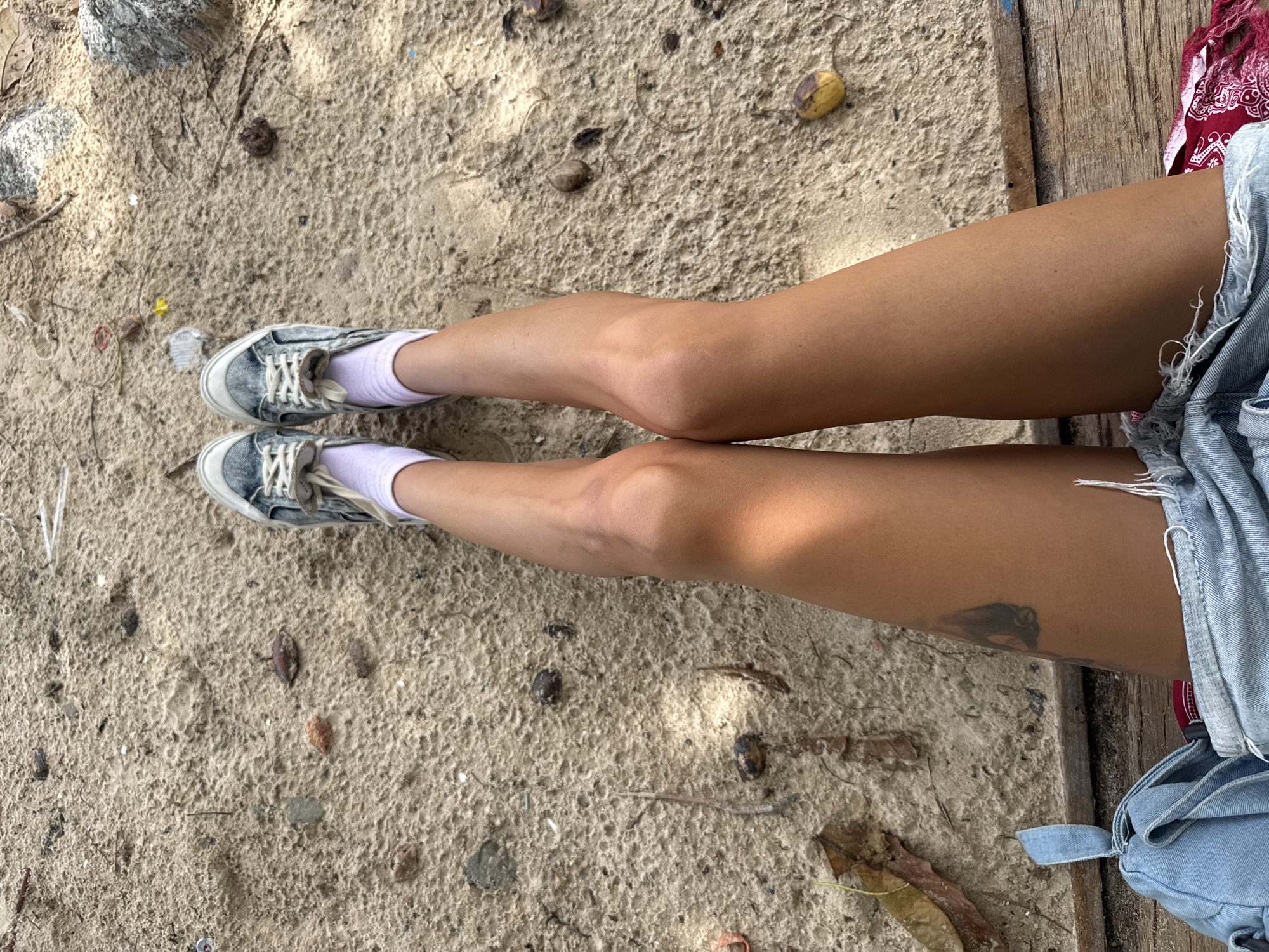 The image shows a person's legs and feet resting on a sandy surface, possibly a beach, in Choeng Thale, Thailand. The person is wearing light-colored sneakers and white socks. Their legs are tanned, and there is a tattoo visible on the left thigh. Denim shorts are worn, with some frayed edges. The sandy ground is littered with small pebbles, dried leaves, and other debris. Wooden planks are visible beneath the sand. The lighting suggests it is daytime, with some shadows cast by unseen objects.