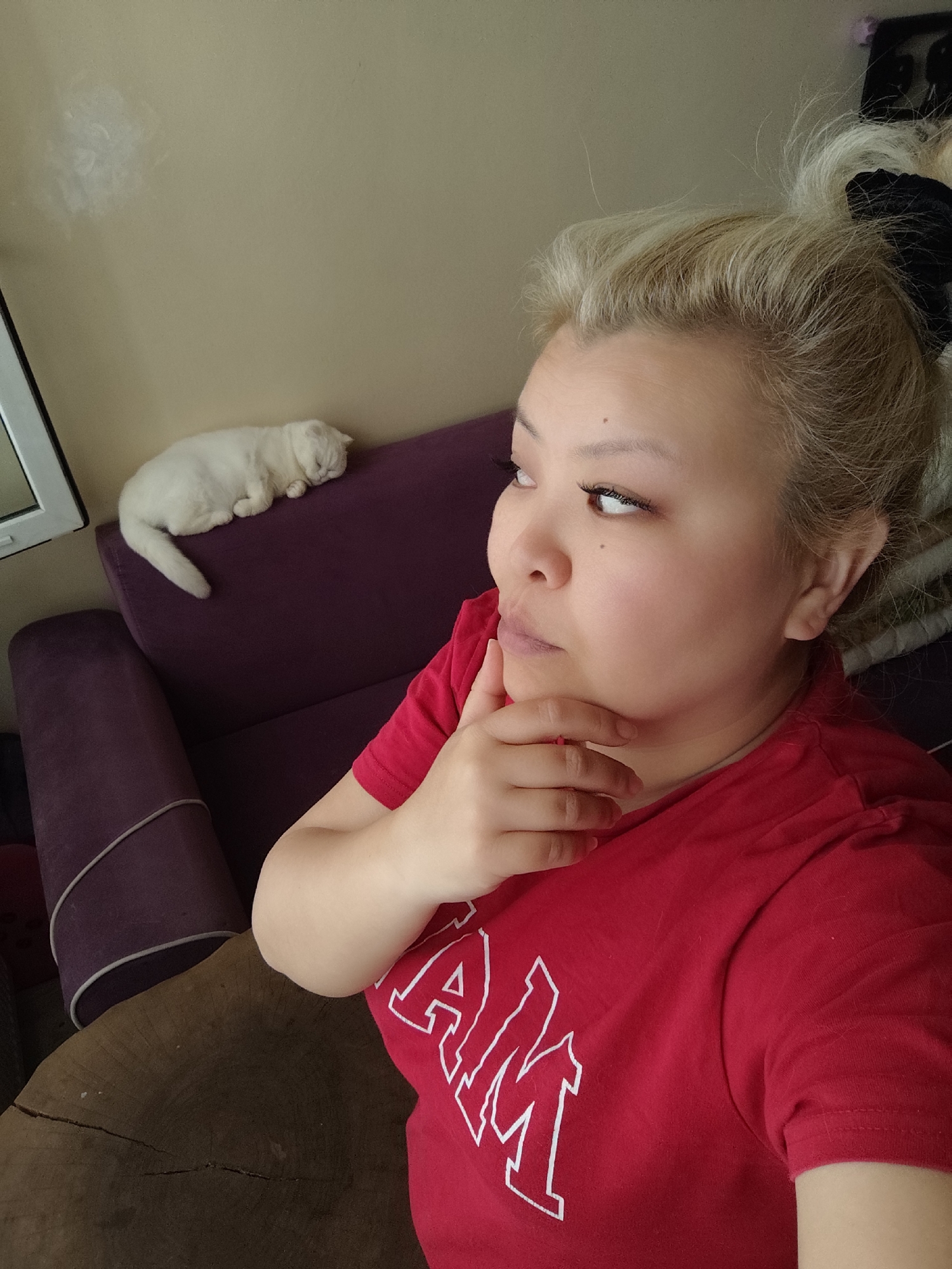 A woman with blonde hair pulled back into a messy bun and wearing a red t-shirt is posing indoors with her hand on her chin, looking to the left. A white cat is sleeping on a purple couch behind her. The location is assumed to be in Sarıyer, Turkey, based on previous information, but there is no explicit evidence of the city in the image. A wooden table is visible in the lower left corner. The wall is a light brown color with a white patch near the upper left corner. A white-framed window is also visible on the left.