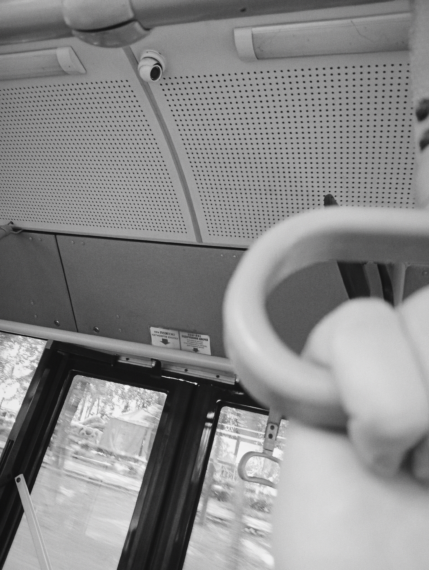 The image shows the interior of a public transportation vehicle, possibly a bus, in the city of Poltava, Ukraine. The view is from the inside, looking towards the ceiling and a window. A security camera is mounted on the perforated ceiling panel. A handle for passengers to hold onto is prominently visible in the foreground, partially out of focus. Through the window, blurred scenery is visible, suggesting the vehicle is in motion. A sign in Ukrainian is near the window.