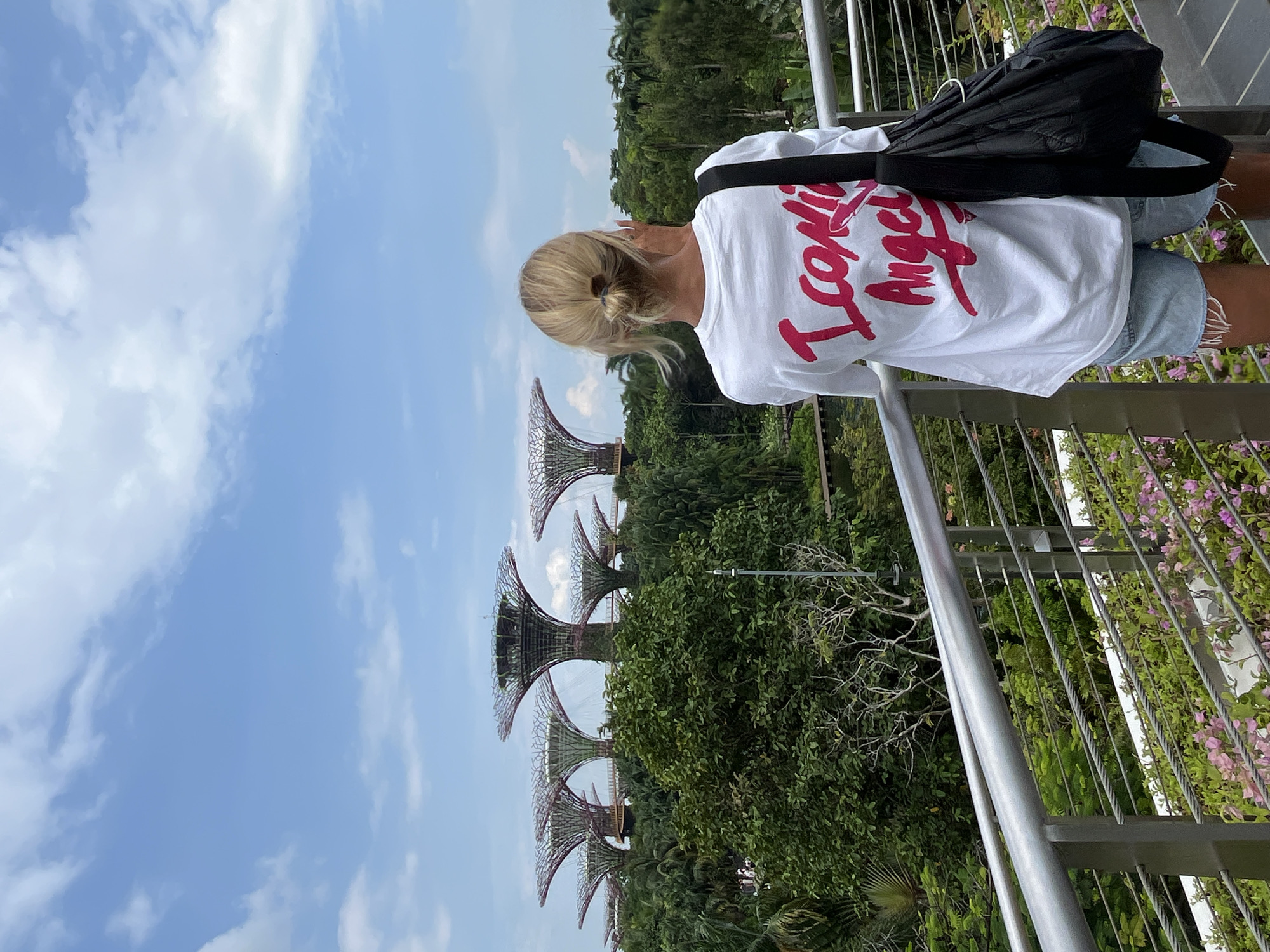 A woman with blonde hair pulled back in a bun stands with her back to the camera, looking out at the Supertree Grove at Gardens by the Bay in Singapore. She is wearing a white t-shirt with a pink graphic design, light blue denim shorts, and a black bag slung over her right shoulder. The Supertrees, iconic vertical gardens of Singapore, are visible in the background amidst lush greenery. The sky is blue with scattered white clouds. She is standing on a walkway with a metal railing, and there are flowering plants in the foreground.