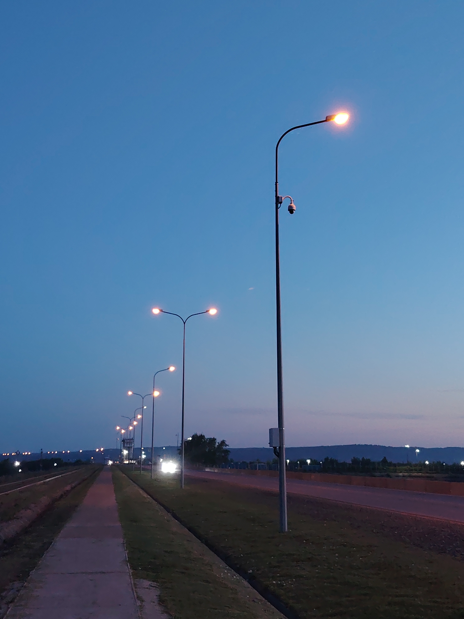 This image depicts a street scene at dusk or early evening in what appears to be the city of Chaikovsky, Russia. The image is taken from the side of a pedestrian walkway with street lamps lining a road or highway. The sky is a gradient of blue. The lamps are illuminated, casting a warm glow. In the distance, car headlights can be seen on the road. Trees and hills are visible on the horizon. One street lamp is equipped with a surveillance camera. The ground is covered with grass along the walkway and the road.