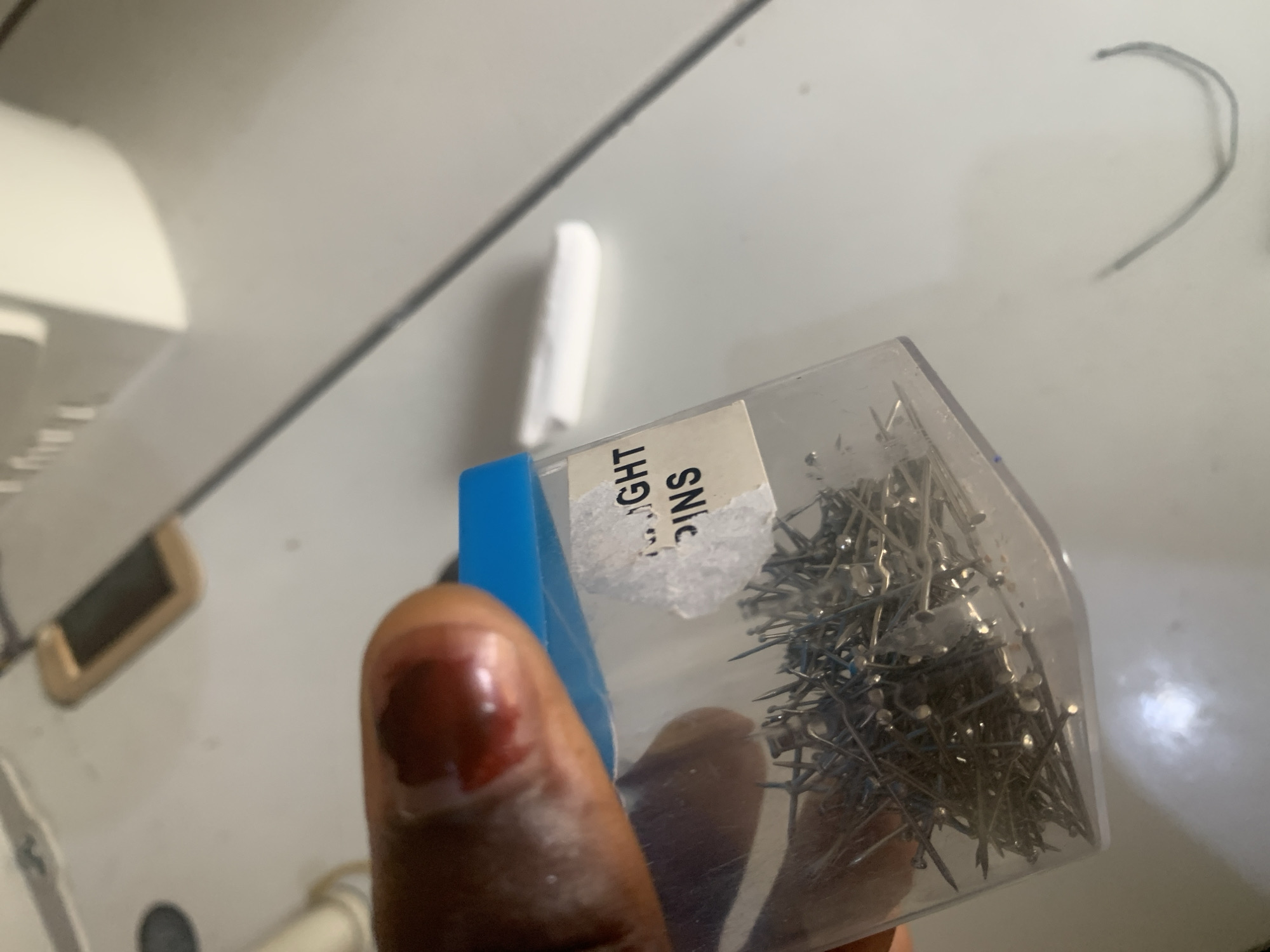 A close-up shot shows a person's finger with a bloody wound held next to a small, clear plastic bag filled with straight pins. A white label is affixed to the bag, partially obscuring the contents, with the words "IGHT" and "PINS" visible. The background suggests an indoor environment with a light-colored, textured surface, possibly a table or workbench, with hints of electronic equipment or appliances. There is no specific indication of the location within Jauro Yinu, Nigeria.