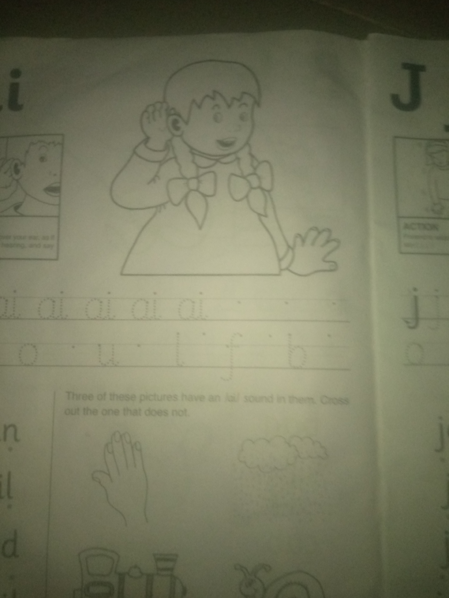 The image depicts a page from a children's activity book, likely focused on early literacy. On the left side, the letter "i" is prominently displayed, followed by the letter "J" on the right side. Below the letter "i" are smaller images and text, including what appears to be a diagram of an ear and the words "over your ear, as if hearing, and say".
The central portion of the page features a large illustration of a girl with pigtails, holding her hand to her ear, suggesting the act of listening. Below this illustration, there are dotted lines with the letter "ai" repeated several times, intended for tracing. Further down, there are dotted outlines of letters: "o", "u", "l", "f", and "b".
Below the tracing section, a sentence reads: "Three of these pictures have an /aɪ/ sound in them. Cross out the one that does not." Following this instruction are three drawn images: a hand, a cloud, and a snail.
Further down the page, more letters are visible, "n", "il", and "d" on the left, and "j" and "o" on the right. At the bottom of the left side, there are outlines of a train and a snail.
The image does not provide any information that would allow for its location to be identified in Jauro Yinu, Nigeria. The scene is entirely consistent with an indoor educational setting.