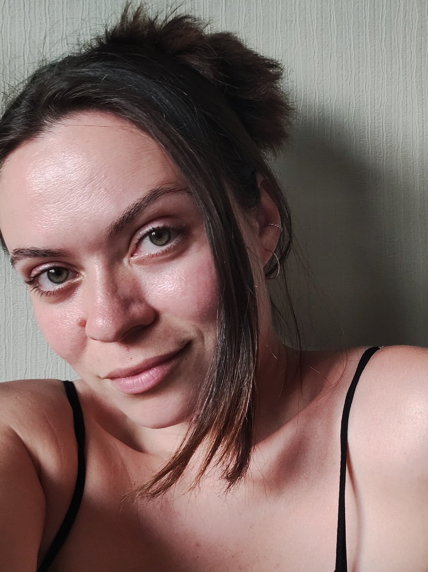 A close-up, eye-level shot depicts a young woman with fair skin and dark brown hair styled in a messy bun. A few strands of hair fall loosely near her right cheek. She has green eyes and a slight smile. Her attire consists of a black spaghetti-strap top. The background appears to be a plain, light-colored wall. There is no indication of the location being in Chaikovsky, Russia.