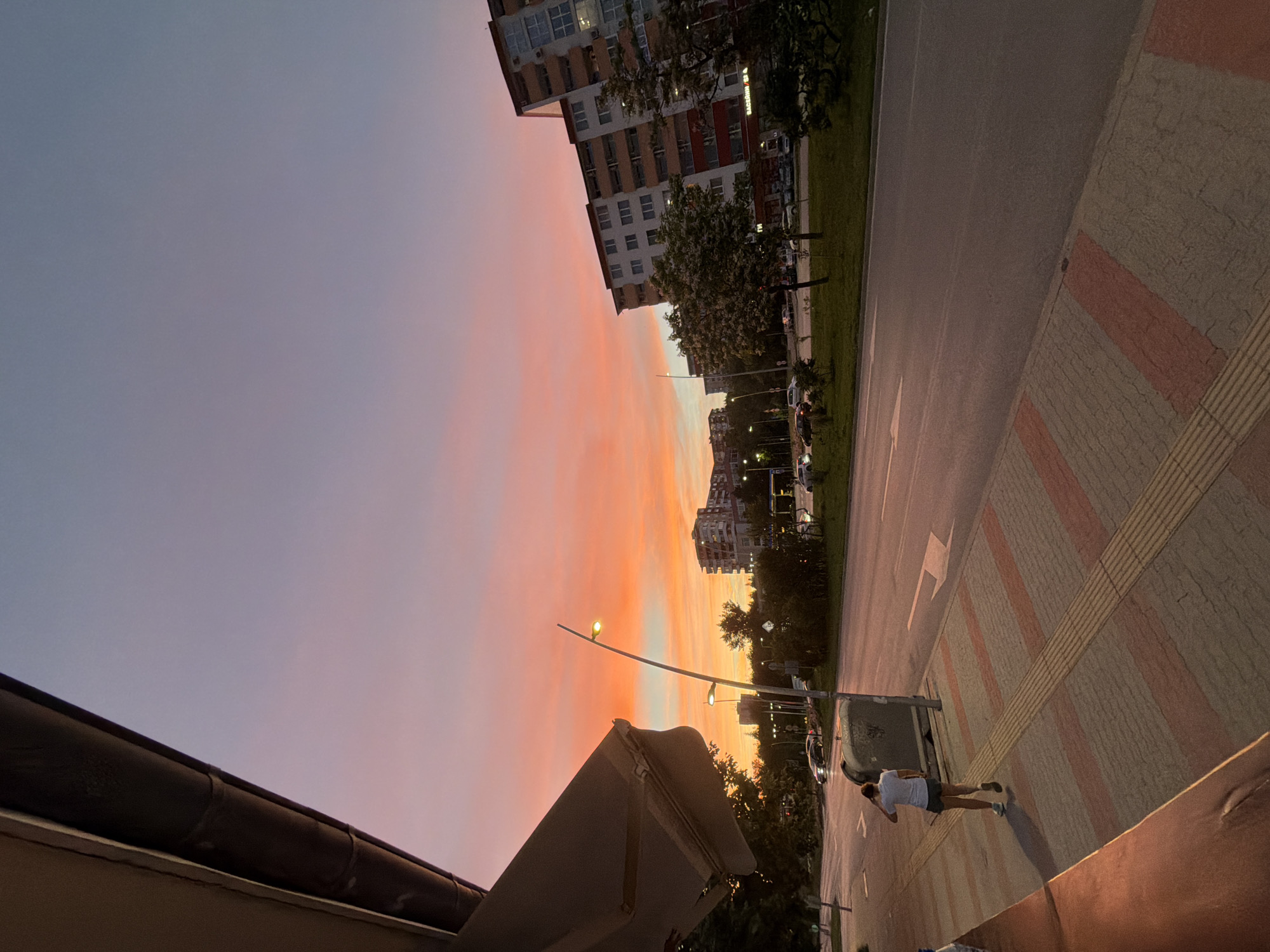 The image depicts a street scene in Veliko Tarnovo, Bulgaria, during a sunset. The sky is a gradient of pink, orange, and blue. On the left, a building overhang with a canvas awning is visible. A person wearing a white t-shirt and dark shorts is walking away from the camera on a sidewalk paved with red and beige rectangular tiles and textured yellow tactile paving. Beside the sidewalk is a large, gray garbage bin. Across the street, several cars are parked and driving, and beyond them are apartment buildings and trees. The road has clear lane markings. The overall lighting suggests it is dusk.