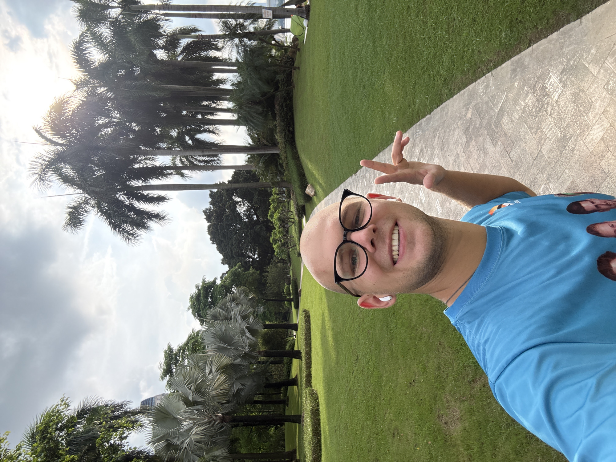 A smiling, bald man with glasses and white earbuds is taking a selfie in a park. He is wearing a bright blue t-shirt. The man is standing on a paved walkway, and to his right is a well-maintained grassy area. Behind him, there are several tall palm trees, and in the distance, some buildings are visible. The sky is cloudy. The location is Haizhu District, China.