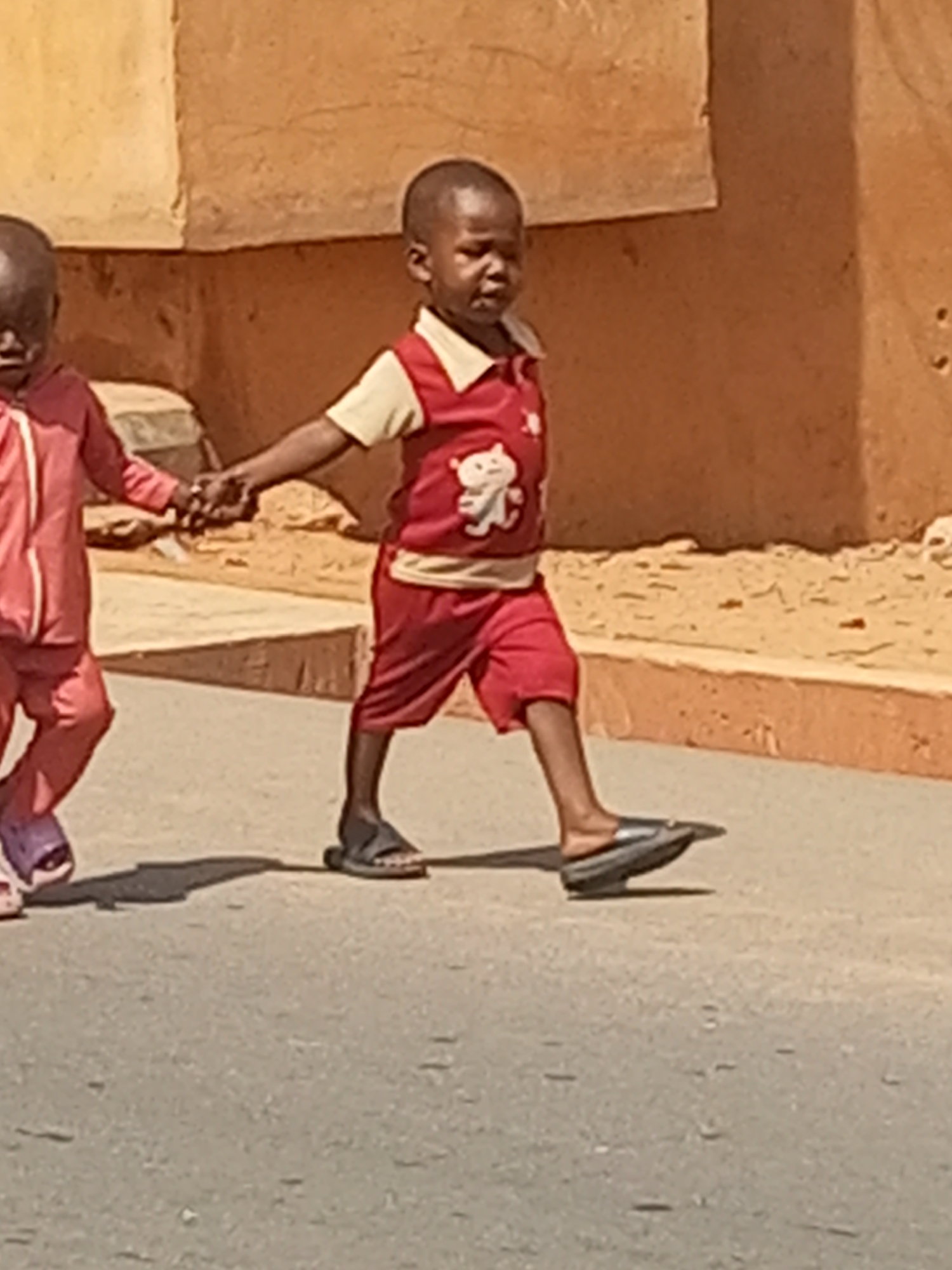 This image depicts two young children, likely boys, walking hand-in-hand along a paved surface in Tempure, Nigeria. The child on the right, more prominently in focus, wears a red vest with a white collar and a white teddy bear-like cartoon character emblem, over a light yellow long-sleeved shirt and red shorts. He has dark sandals on his feet, one appearing black and the other dark grey. His expression is neutral. The second child, partially visible and blurred on the left, wears a red full tracksuit or jumpsuit and light purple sandals.
The background features a light brown building, likely constructed from mud or plaster, with visible architectural textures and ledges. Strong, defined shadows cast by the children suggest bright, clear daylight, indicating a sunny day, possibly mid-morning or afternoon. The setting appears to be a local street or pathway. No visible text is present in the image. The scene portrays an everyday moment of companionship between the two children.