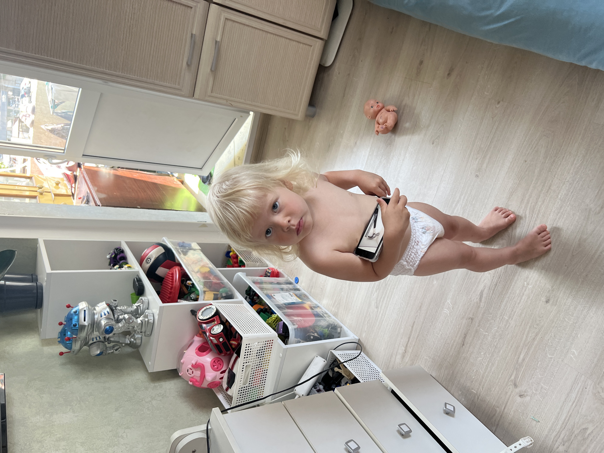 The image shows a young child, likely a toddler, with blonde hair and blue eyes, wearing a diaper. The child is sitting on a light-colored wooden floor. In their right hand, they are holding a smartphone. In the background, there is a white shelving unit filled with various toys, including toy cars, a robot toy, and a pink pig toy. Above the shelving unit, there are light-colored cabinets. To the right of the child, there is a small doll lying on the floor and a blue blanket. The overall scene suggests a playroom or a child's bedroom. The lighting appears to be natural, indicating it could be daytime. No specific text is visible in the image. The context suggests the location is Sochi, Russia.