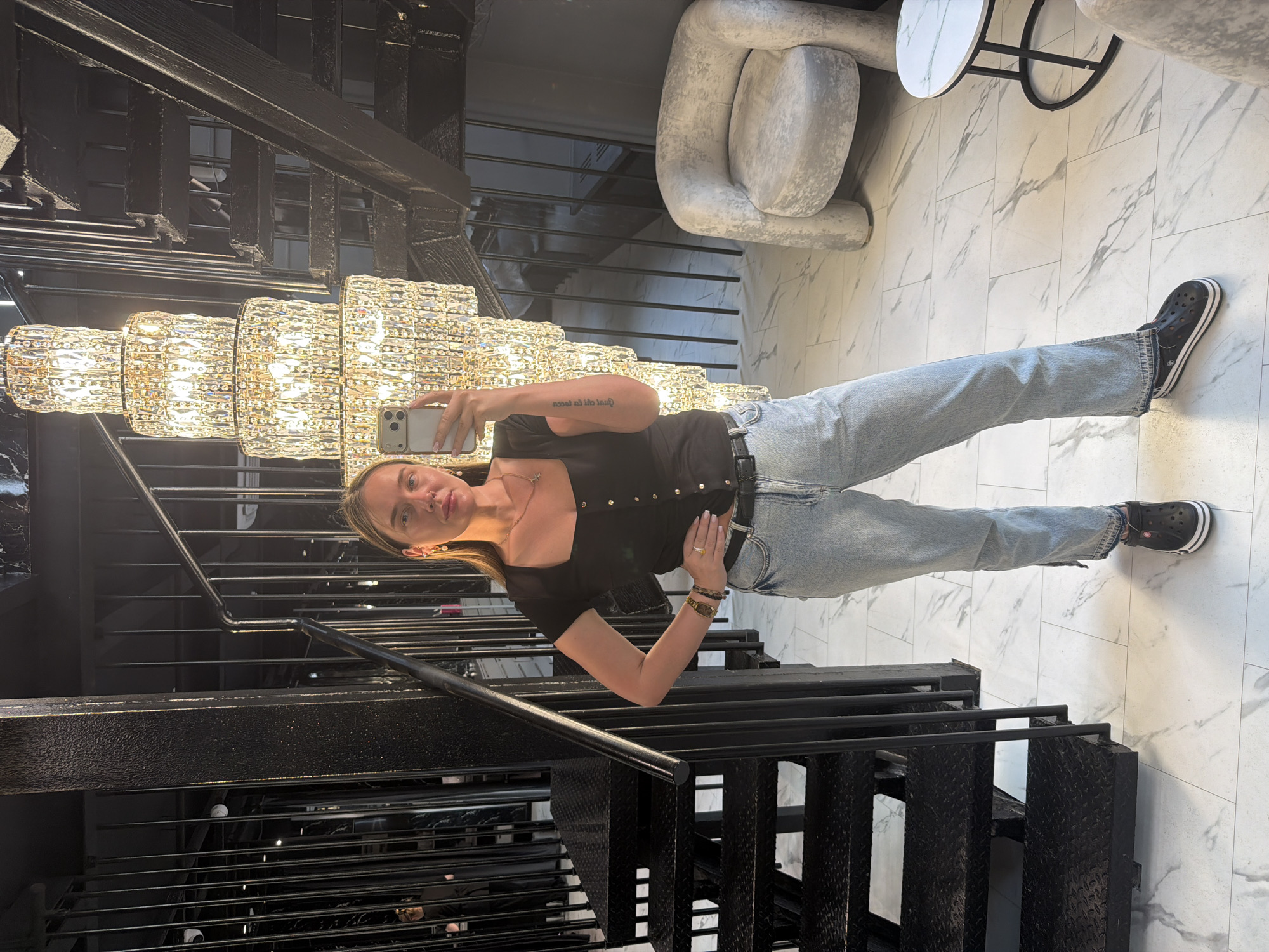 A woman is captured in a mirror selfie inside a modern building in Укромновское сельское поселение, Ukraine. She stands centrally, holding a light-colored smartphone with her right hand to take the photo, partially obscuring her face. Her left hand rests on her hip. She wears a black square-neck top, light blue jeans with ankle slits, a black belt, and black Crocs with white striped soles. Accessories include a watch and rings on her left hand, a thin gold chain with a cross pendant, and light-colored drop earrings. A tattoo is visible on her left forearm.
The background features a grand, multi-tiered crystal chandelier illuminating the scene with warm light, positioned behind and to the right of the woman. To her left, a prominent black metal staircase with textured steps and vertical railings ascends. The floor is covered in white marble-patterned tiles. On the right side of the image, a light gray, textured armchair with a rounded design and a small round side table with a marble top and black base are visible. Dark walls and additional vertical black metal architectural elements create a structured background. A faint reflection of another individual is discernible in the dark background behind the staircase on the far left.