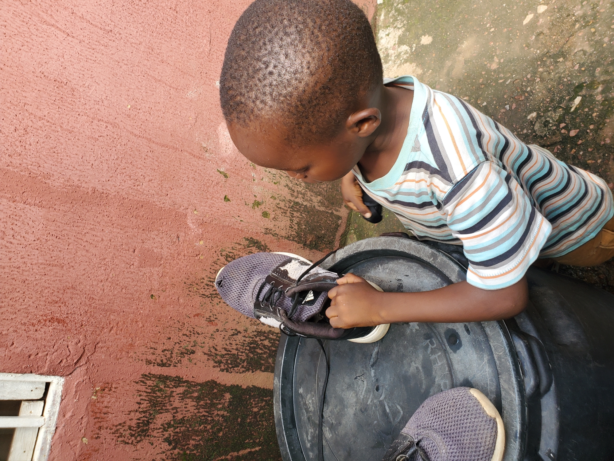 A young Black boy, likely under the age of five, is seen from a high angle, looking down at a pair of grey sneakers placed on top of a large, black plastic drum. He is wearing a striped t-shirt with horizontal bands of light blue, white, grey, and orange. His skin is dark, and he has short, dark hair. The boy is holding one of the sneakers with his right hand, seemingly examining it.
The scene appears to be outdoors, with a textured, reddish-brown wall on the left side of the frame. A white wooden window frame is visible near the bottom left corner. The ground is a mix of concrete and dirt, with some patches of moss or dark growth. The lighting suggests it is daytime. The overall setting seems to be a somewhat unkempt outdoor area, possibly a yard or courtyard in a residential area. The focus is on the boy and his interaction with the shoes. There is no discernible text in the image.