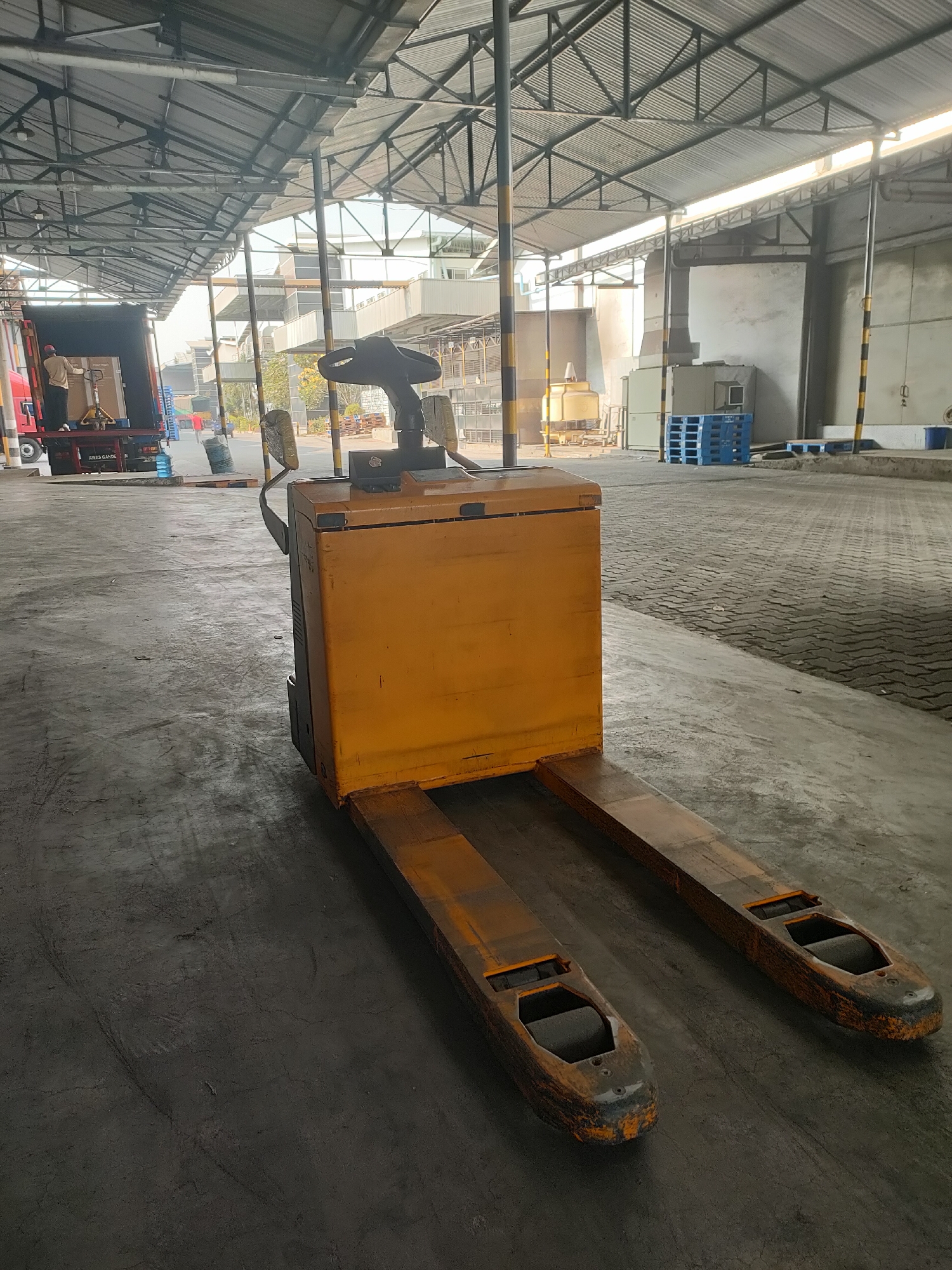 The image depicts an industrial warehouse or loading dock area in Sayung, Indonesia. In the foreground, a yellow manual pallet jack is parked on a concrete floor. The pallet jack has two forks extending forward. The background shows a large, open-sided warehouse structure with a corrugated metal roof supported by metal beams and columns. To the left, a red truck is parked, with a person visible near the back of the truck, appearing to be involved in loading or unloading. Blue plastic pallets are stacked in the middle ground. The overall environment suggests a logistics or distribution facility.