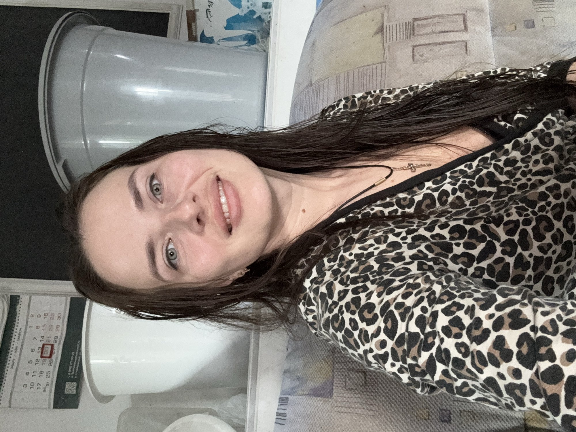 This is a portrait of a young woman with long, dark hair, smiling at the camera. She is wearing a leopard-print top. A small gold cross necklace is visible around her neck. In the background, to the left, is a large, grey, cylindrical object, possibly a bucket or a container. To the right, a textured surface, possibly a sofa or a wall covering, is visible. A calendar is partially visible on the left side of the image. The calendar shows the month of March and the dates are in Russian. The date "20" is circled in red. The lighting suggests it could be daytime, but there are no specific windows or outdoor cues to confirm this. The overall setting appears to be indoors.
