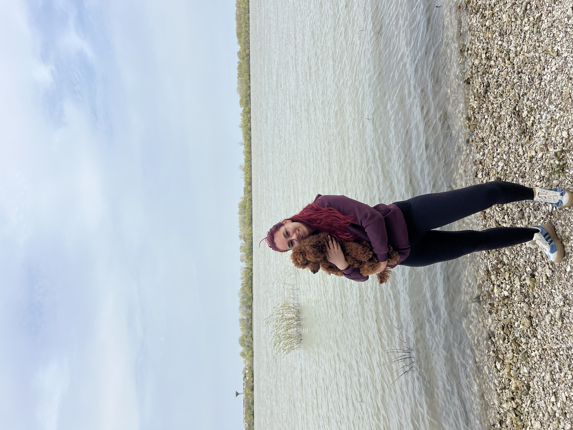 A woman with red hair stands on a gravel beach next to a body of water, holding a brown poodle. The woman wears glasses, a burgundy hoodie, and black leggings, and blue and white sneakers. The gravel beach transitions into a vast expanse of water, likely a river or lake, with the opposite shore lined with trees. The sky above is overcast with a light blue tint. The image is set in Nikopol, Bulgaria.
