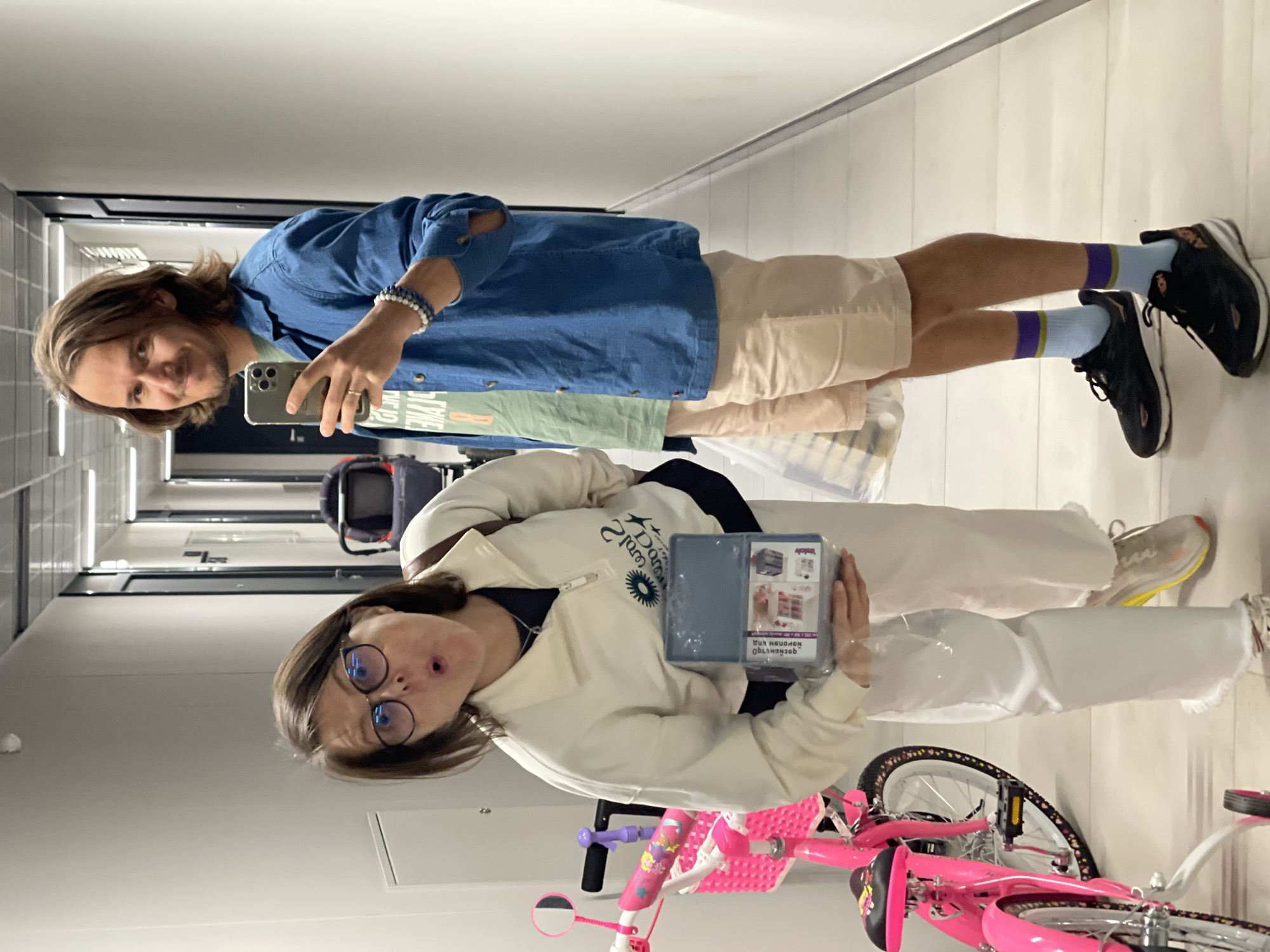 The image depicts a man and a woman standing in an indoor space, possibly a hallway, in the city of Unknown, Unknown. The man, positioned behind the woman, is taking a selfie with his smartphone. He has long hair, wears a blue button-down shirt, beige shorts, and blue socks with purple stripes, and black sneakers. He is also wearing a bracelet. The woman, in front of the man, is holding a packaged product. She is wearing glasses, a cream-colored sweatshirt, white pants, and off-white sneakers. A pink bicycle with training wheels is visible near her. The hallway has light-colored walls and a dark-colored trim along the ceiling. Overhead lights are visible in the ceiling.