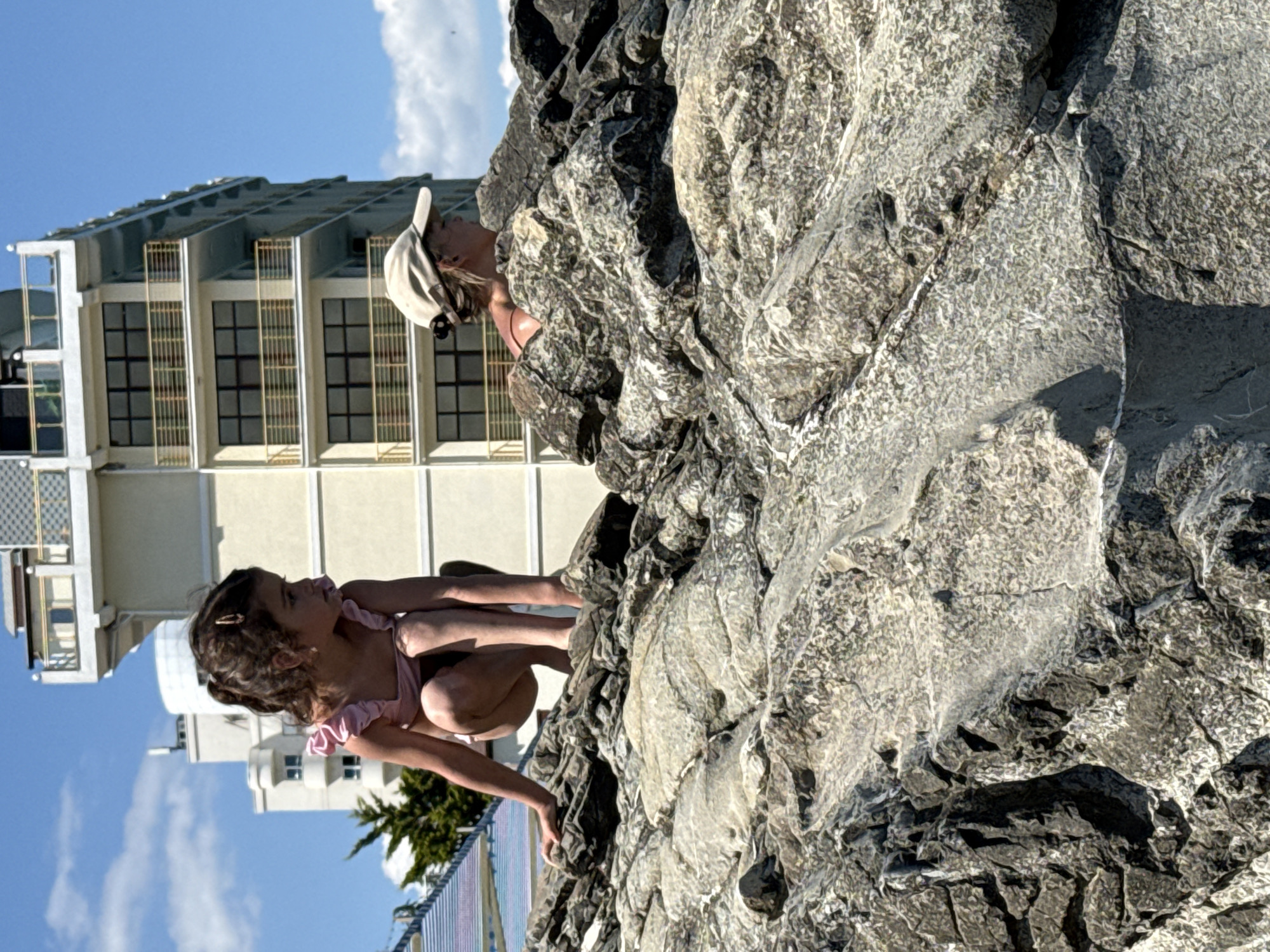 A young girl in a pink swimsuit is crouched on large rocks. She is looking to her right. Behind her, another person wearing a baseball cap is partially visible, also looking to the right. In the background, there is a multi-story building with balconies and windows. The sky is blue with a few clouds. The environment appears to be a coastal area with rocky terrain.