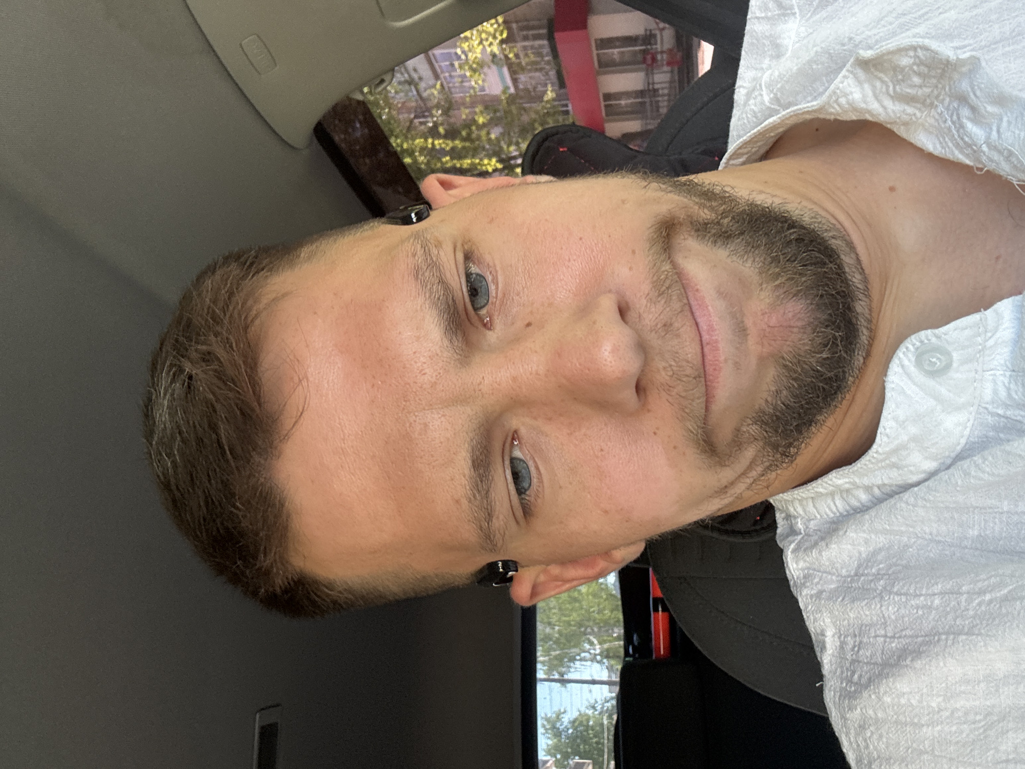 A close-up, indoor shot depicts a fair-skinned man with short brown hair, a beard, and blue eyes. He is wearing what appears to be a white, textured shirt with buttons. He has earbuds in his ears. The image appears to have been taken in a car, as part of the interior of a vehicle is visible, including the ceiling and a window showing a view of trees and a building exterior. The location in the city is Unknown, Unknown.