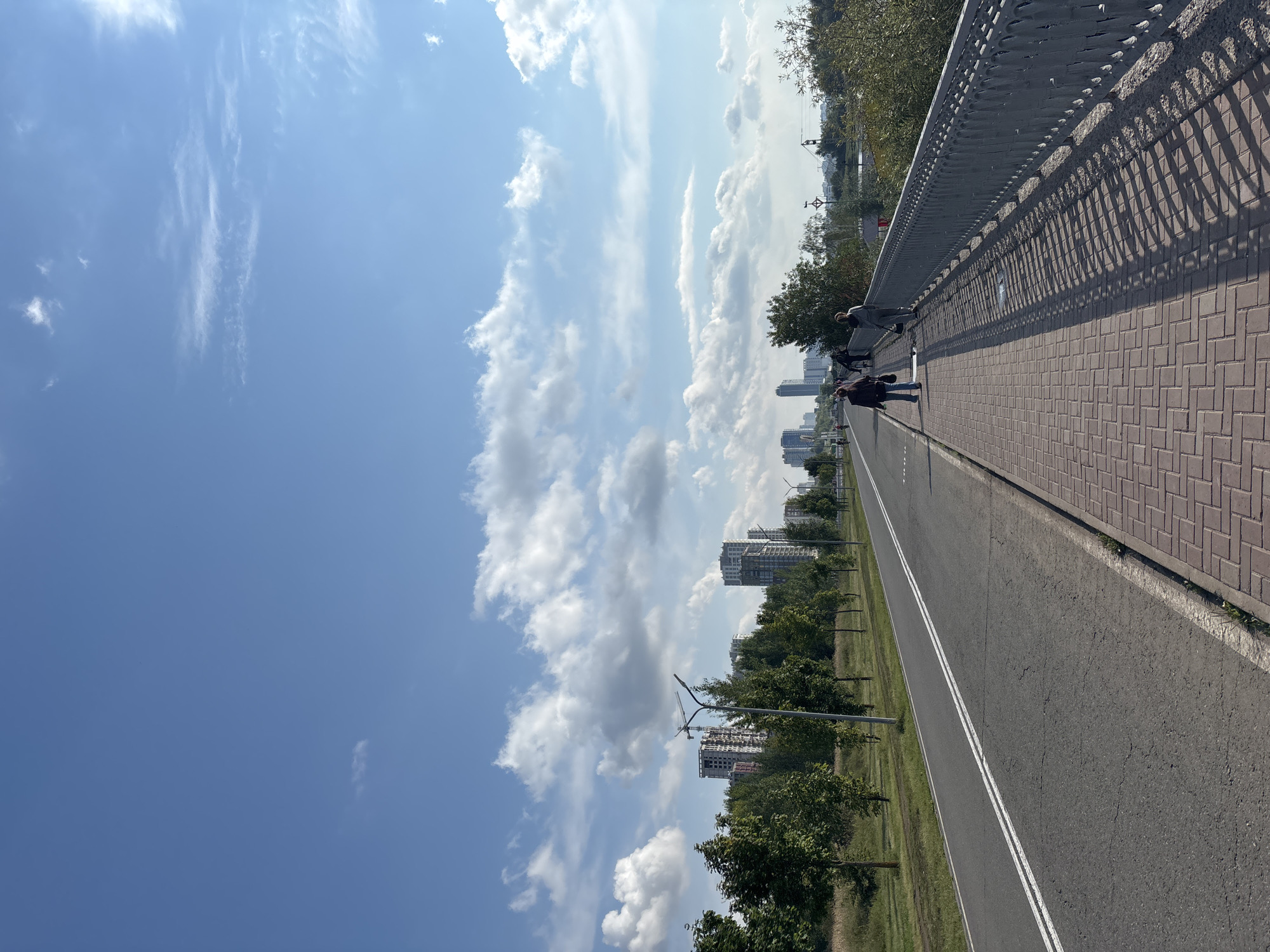 The image depicts a view from a pedestrian bridge overlooking a multi-lane road and a sidewalk in Naberezhnye Chelny, Russia. Tall, modern buildings and construction sites are visible in the distance, surrounded by lush green trees. The sky is a clear blue, dotted with fluffy white clouds, suggesting fair weather. A metal railing lines the right side of the bridge, casting long shadows across the brick-patterned sidewalk. Several individuals are seen walking and cycling on the sidewalk and road, engaged in their daily activities. The overall scene conveys a sense of urban life intertwined with nature, under a bright, sunny sky.