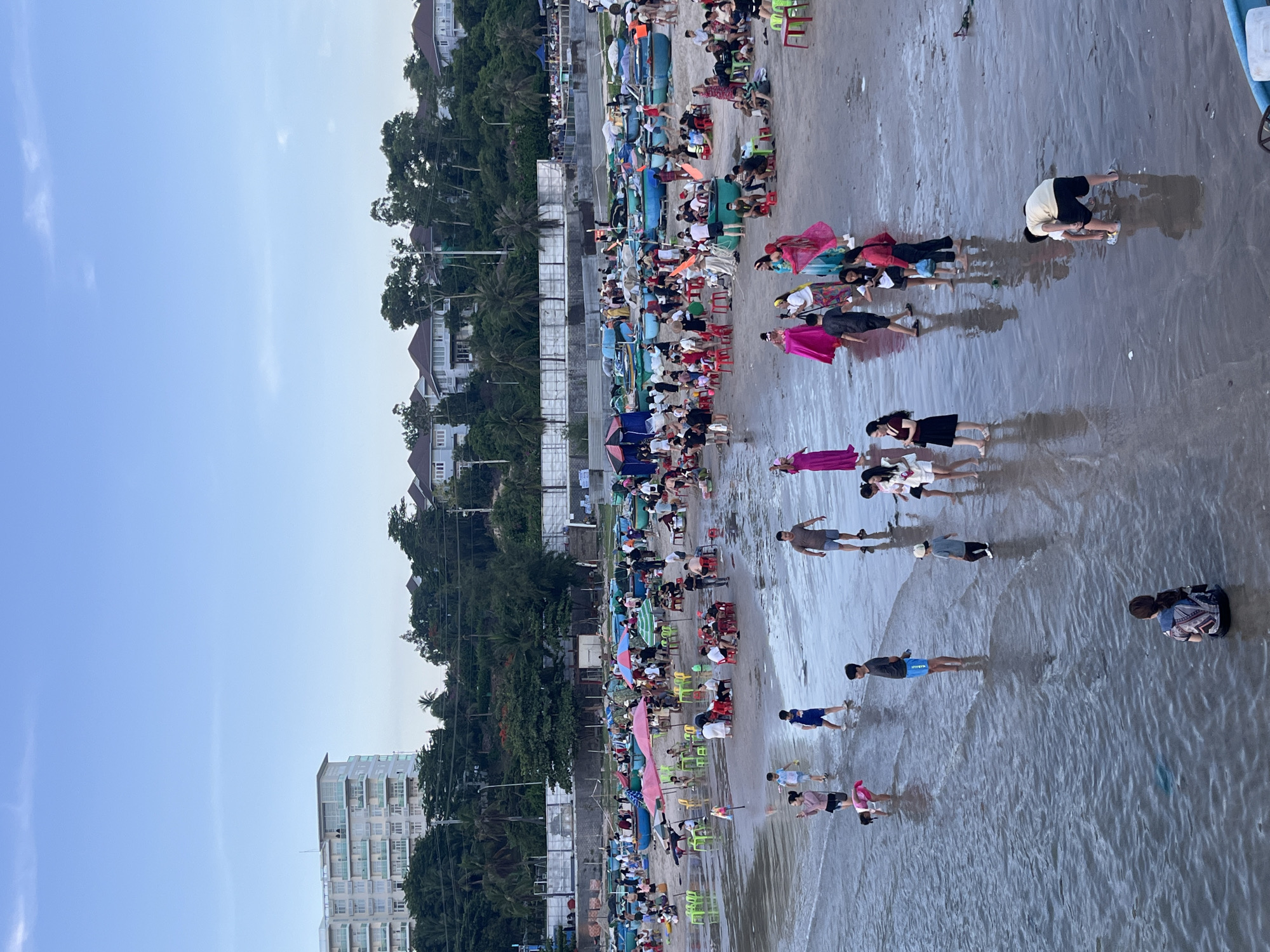This is a wide-angle, eye-level shot of a bustling beach scene in Phu Thuy Ward, Vietnam. The foreground shows the shallow, wet sand with gentle waves lapping onto the shore. Several individuals and groups are scattered across the beach, some wading in the water, others walking, and some engaged in activities on the sand. In the midground, a large number of people are gathered around small tables and chairs set up on the wet sand, suggesting a dining or seating area. Colorful umbrellas provide shade over some of these areas. Numerous small, blue fishing boats are pulled up onto the sand behind this gathering. A variety of people, dressed in casual beachwear, are seen socializing, walking, and interacting. Further in the background, a line of trees and vegetation separates the beach from a row of residential buildings and a larger, multi-story hotel. The sky above is a pale blue with some wispy clouds. The overall impression is one of a lively and popular recreational area.