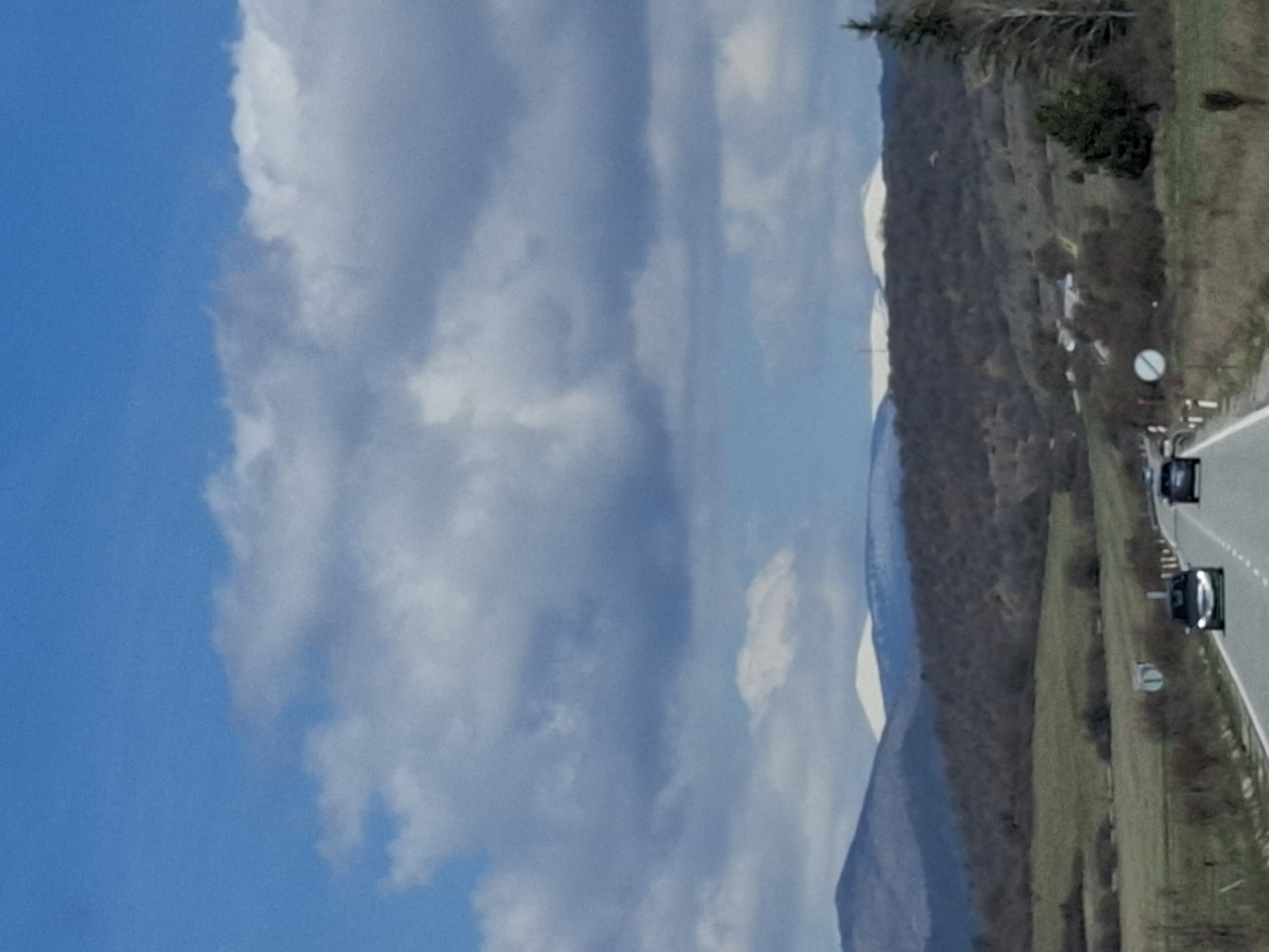 The image shows a daytime scene near Drenovo, Bulgaria. The left portion of the image is dominated by a bright blue sky with large, fluffy white clouds. To the right of the sky and clouds, a mountain range stretches into the distance. On the lower right, a highway is visible with at least two cars traveling along it. The landscape along the highway consists of grassy areas, trees, and shrubs.