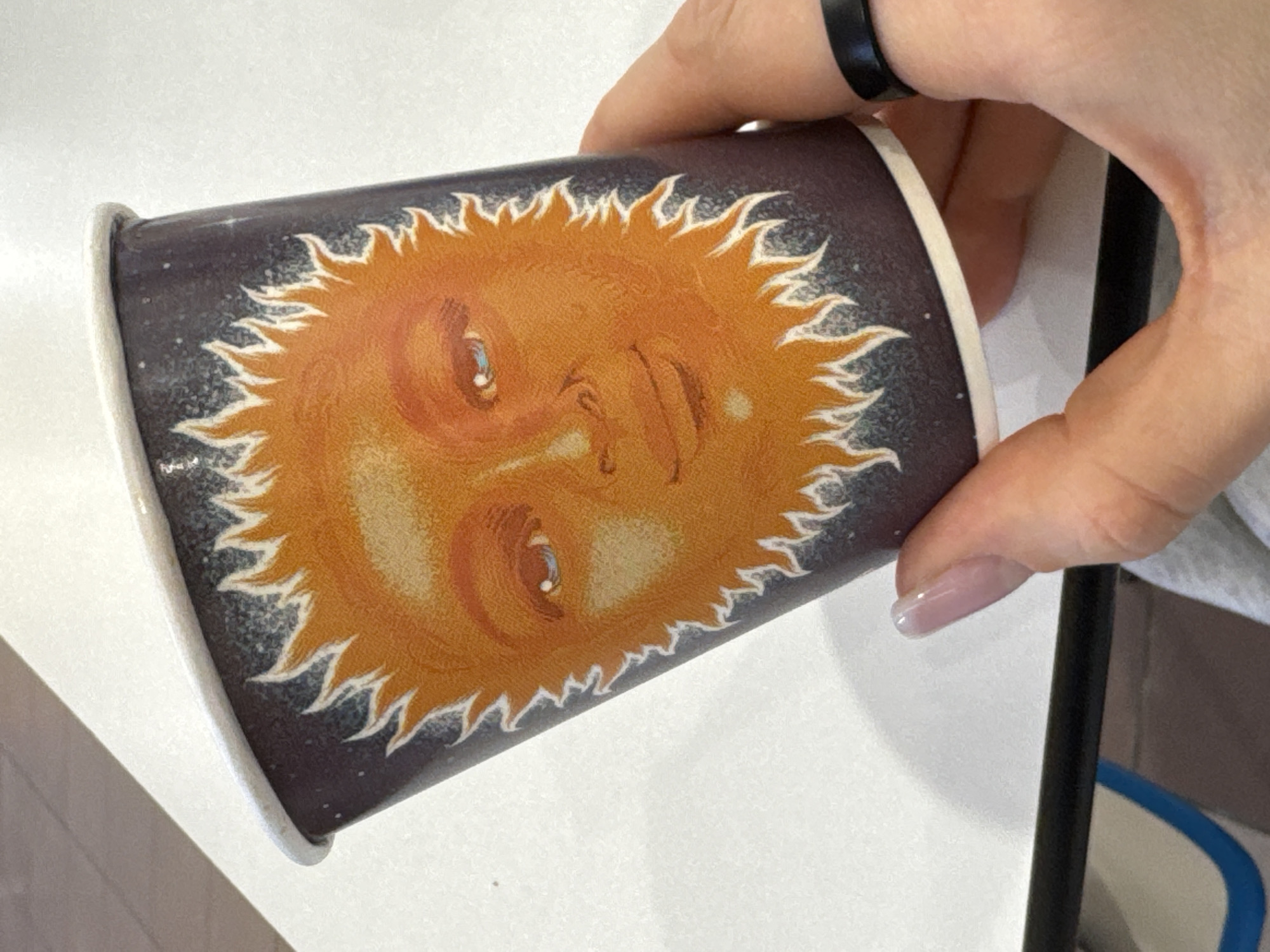A close-up shot depicts a hand holding a paper cup. The cup features a design of a sun-like face with rays emanating from it, set against a dark, star-speckled background. The hand has a black ring on one finger and a light pink nail polish on the fingernails. The background is blurred. No location information or details about the city of Unknown, Unknown, can be derived from this image.